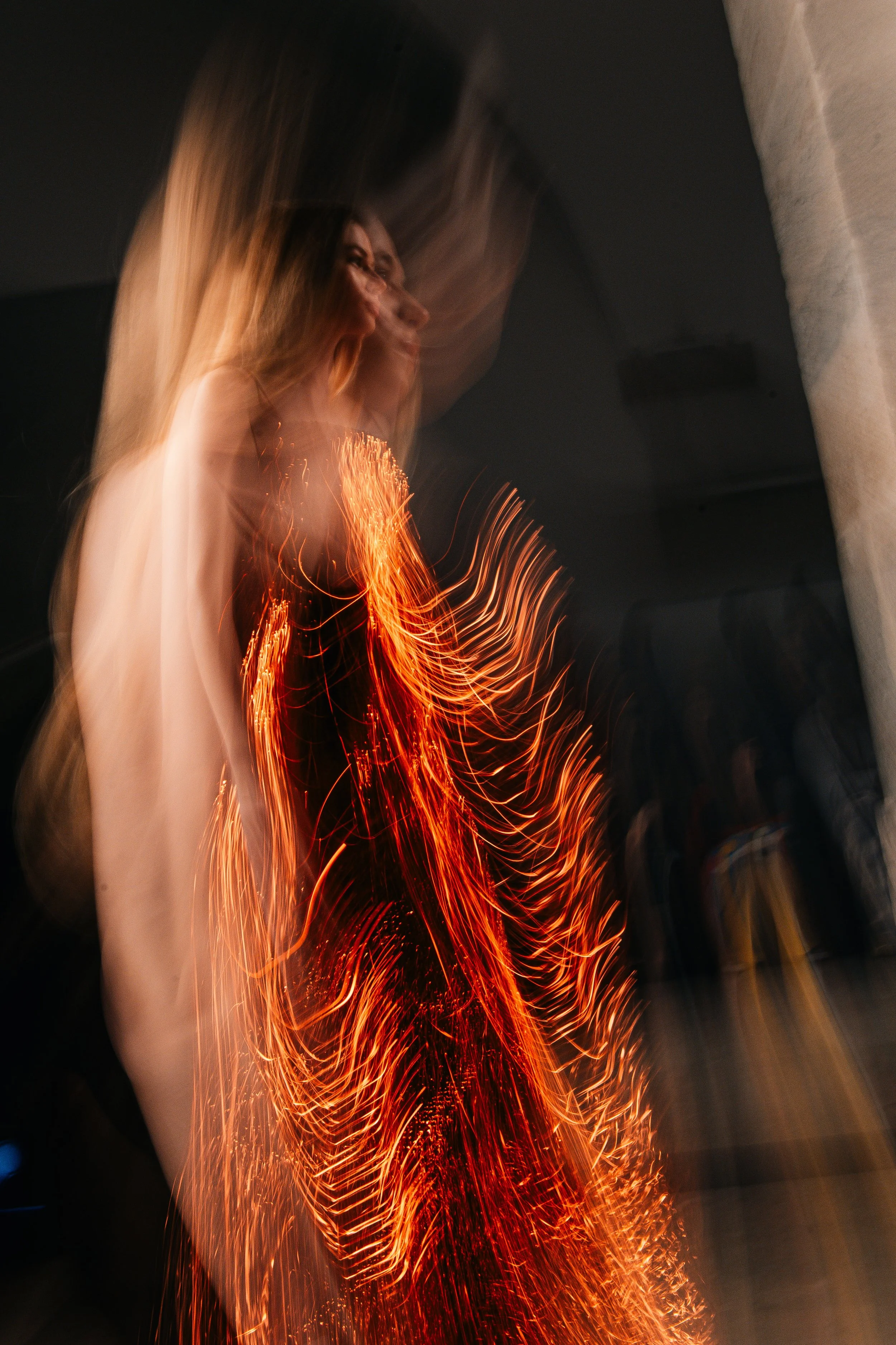 Pessoa com cabelo longo, usando vestido preto, em uma performance com faíscas e luzes vibrantes, movimento e efeitos de luz.