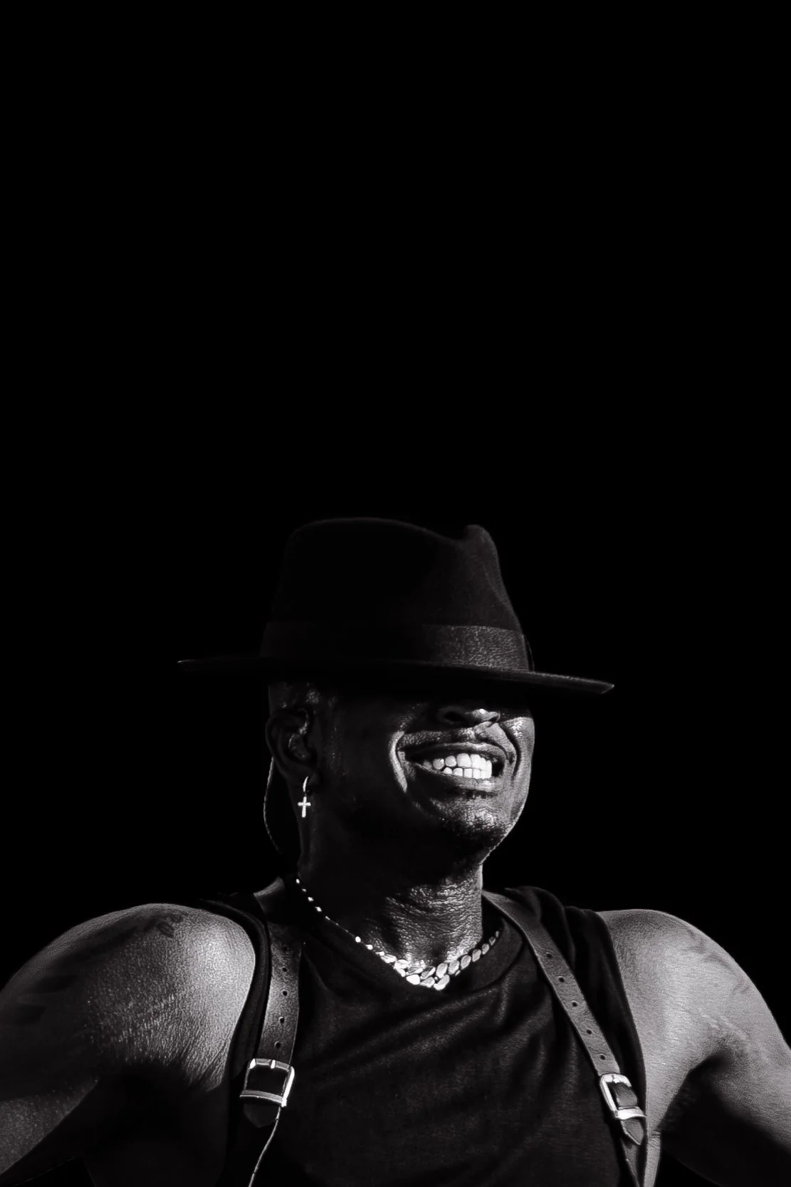 Homem sorridente com chapéu de aba larga, brincos e colar, usando uma roupa sem mangas, em um fundo preto.