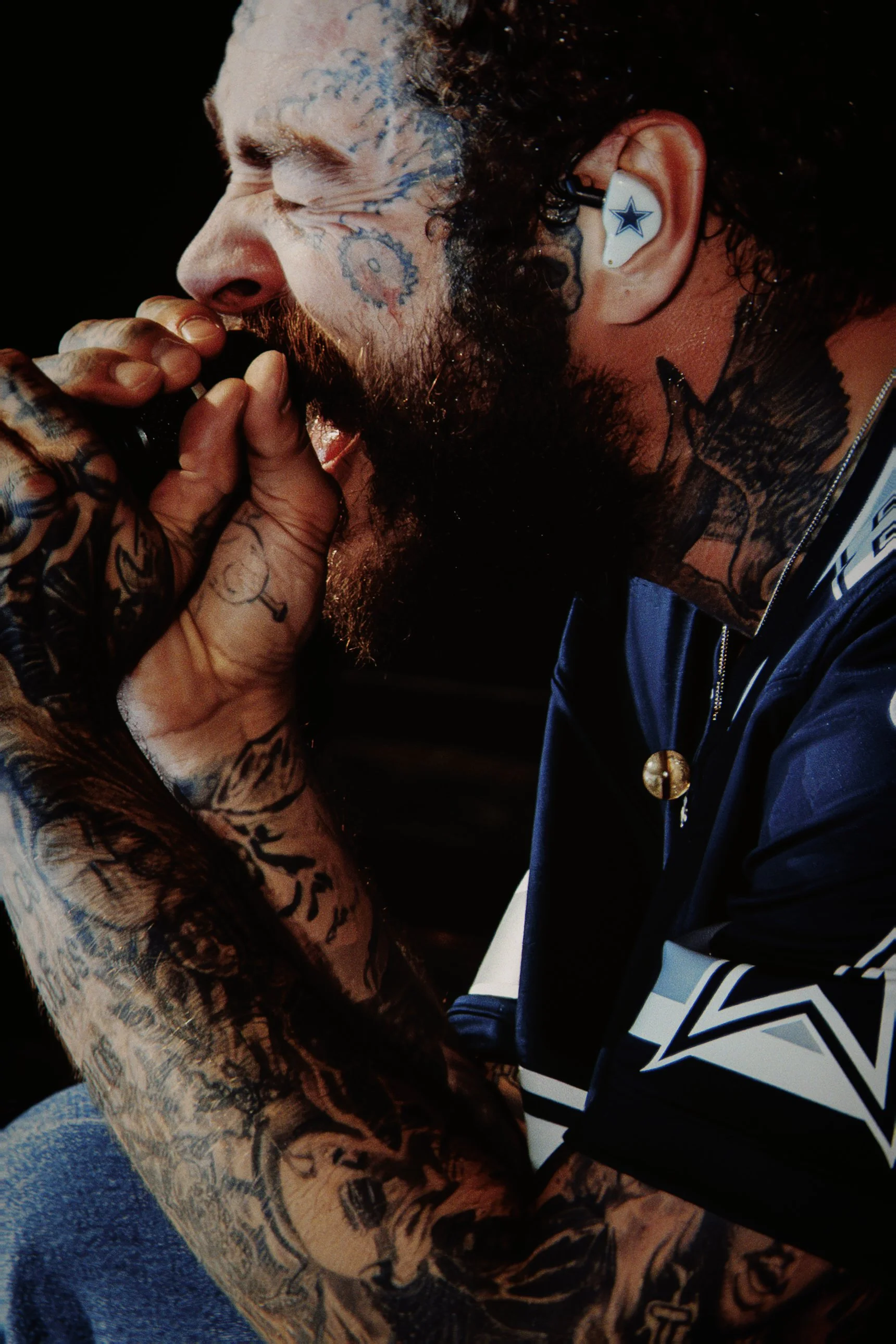 Homem com barba e tatuagens visíveis na cabeça, pescoço e braço, usando fone de ouvido com a estrela do Dallas Cowboys, segurando microfone, em uma apresentação ou performance.