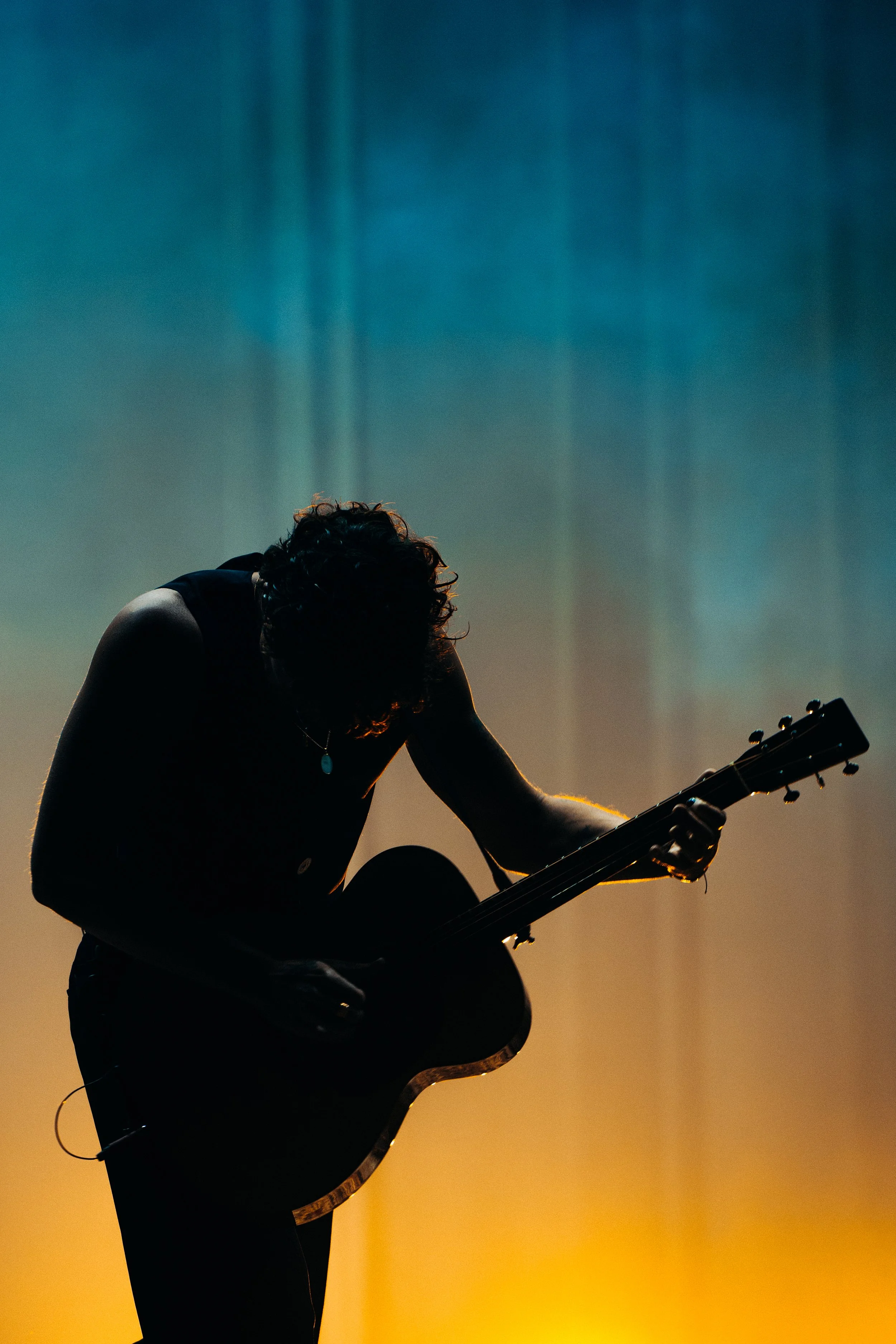 Pessoa tocando violão em um palco com iluminação de fundo em tons de azul e amarelo, aparência de um momento artístico ao pôr do sol ou com iluminação dramática.