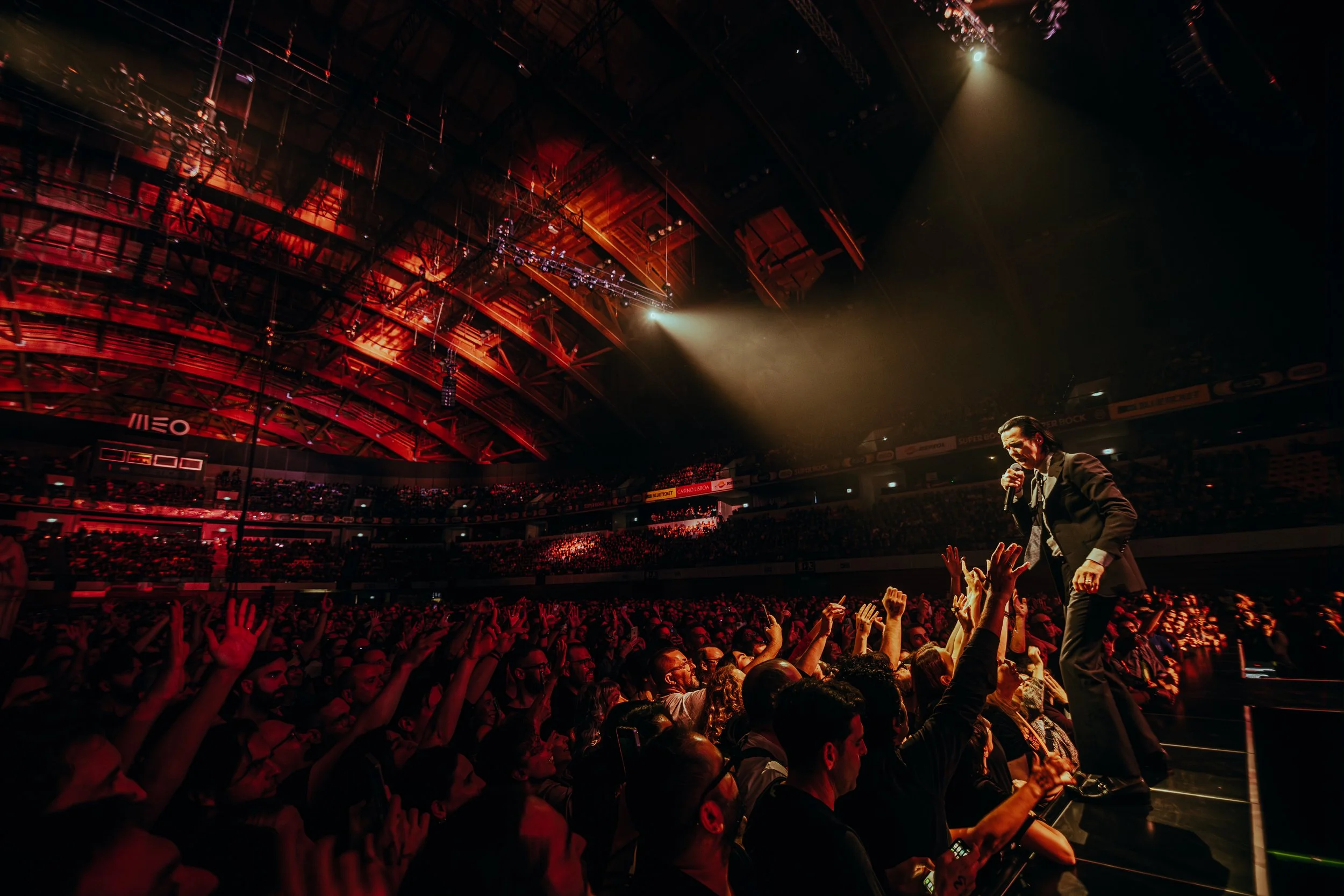 Cena de um show ou evento ao vivo no interior de uma arena com teto avermelhado, iluminação dramática e uma multidão de pessoas com mãos levantadas, ouvindo um homem de terno cantando ou falando ao microfone.