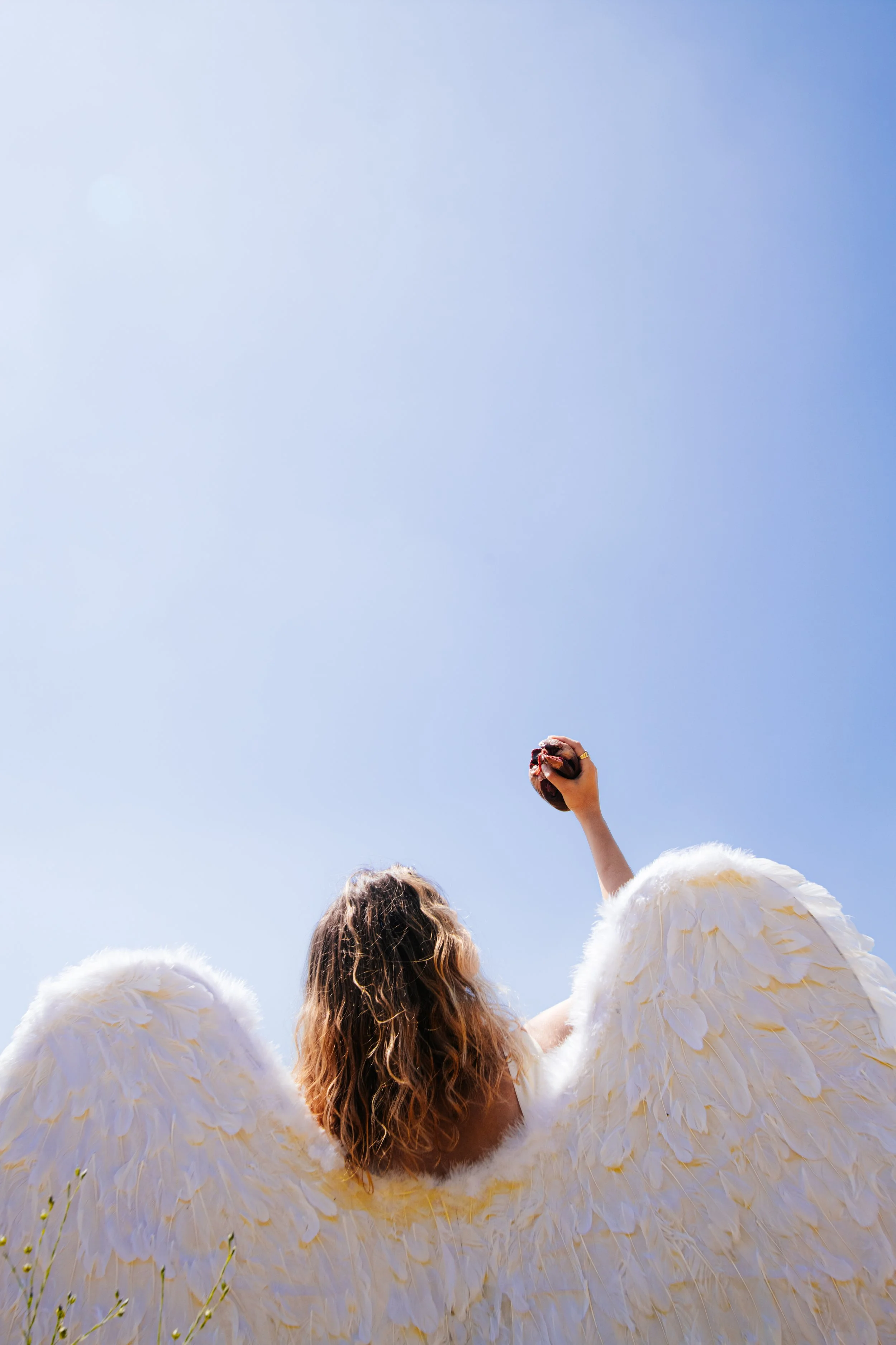 Mulher com asas de anjo segurando um pão na mão, vista de trás, céu azul ao fundo.