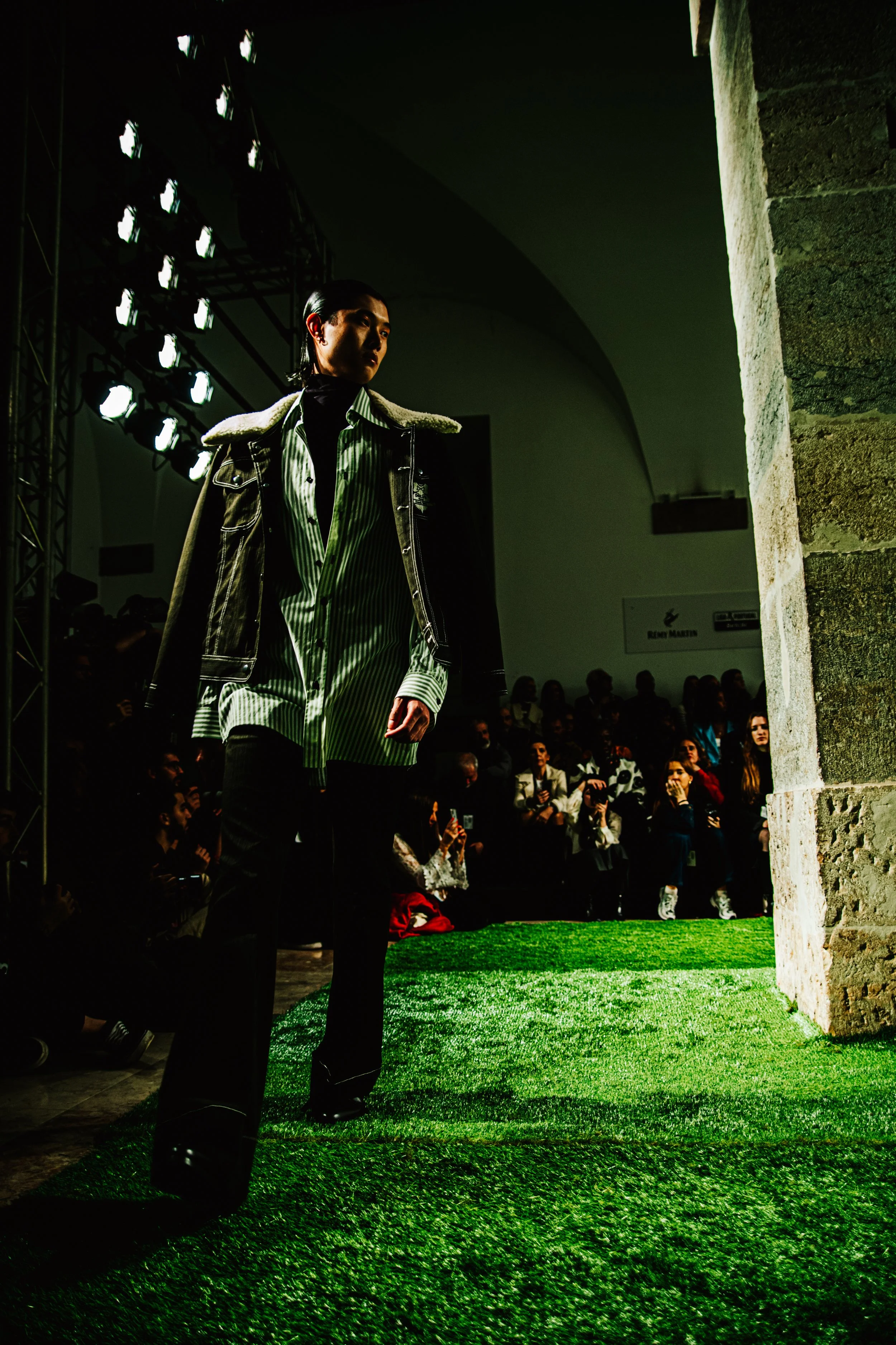 Pessoa andrógina usando jaqueta de couro padrão xadrez em desfile de modas, com iluminação forte de palco e público ao fundo.