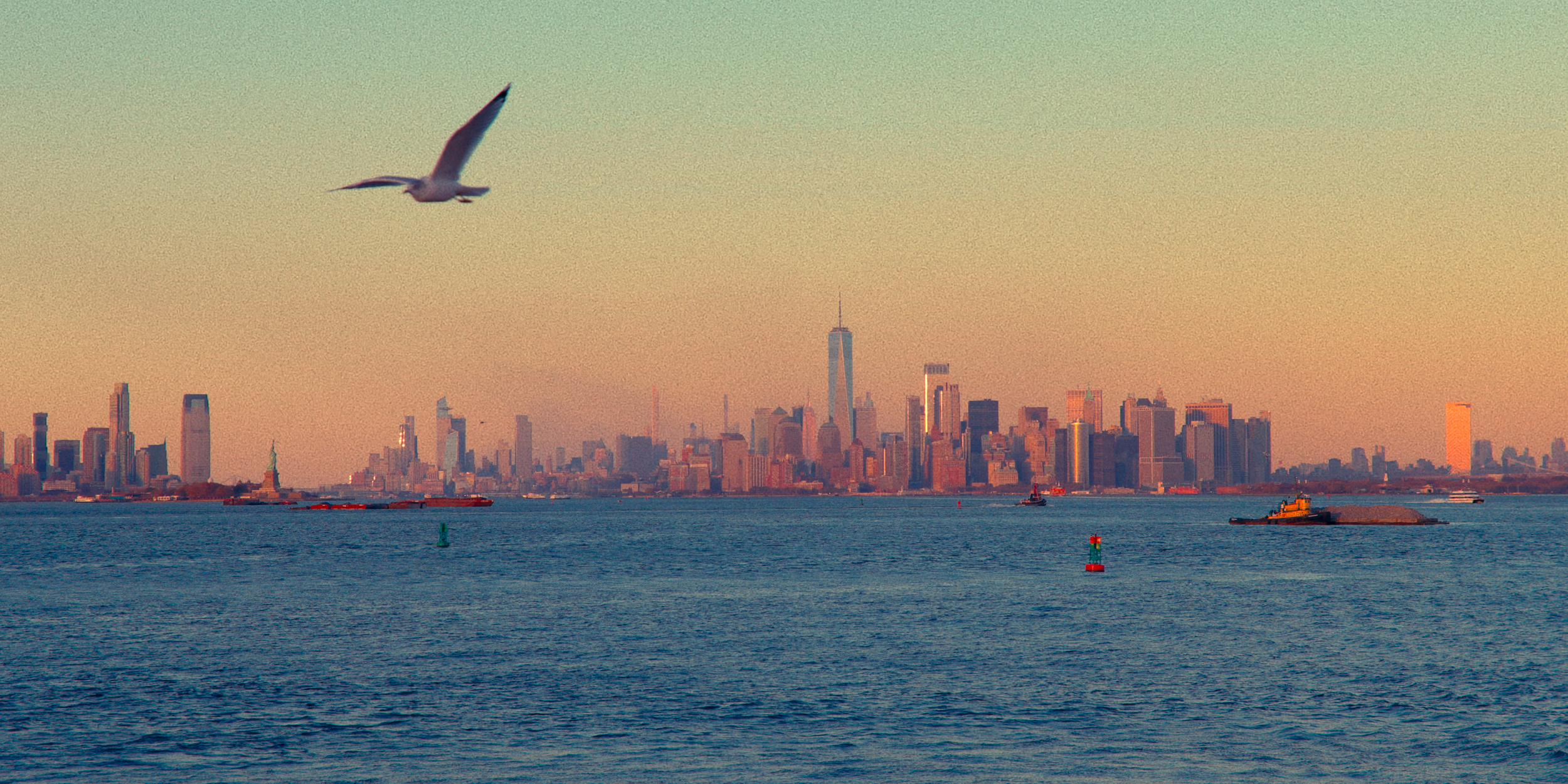 Paisagem de horizonte urbano de Nova York vista da água com várias embarcações e uma gaivota no céu ao pôr do sol.