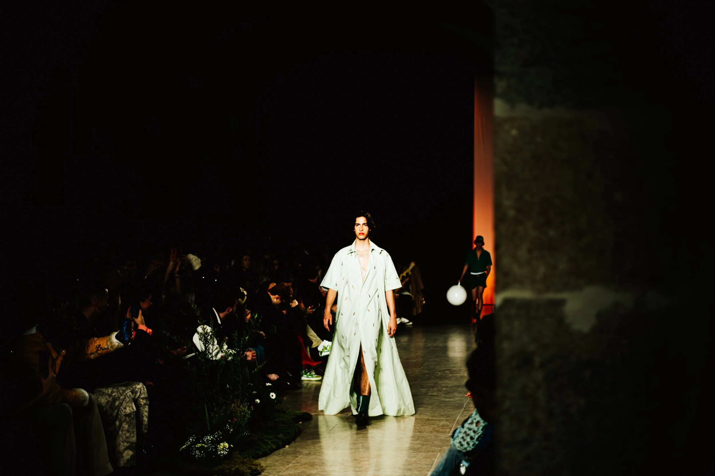 Modelo caminhando na passarela durante um desfile de moda à noite, com público assistindo ao lado.