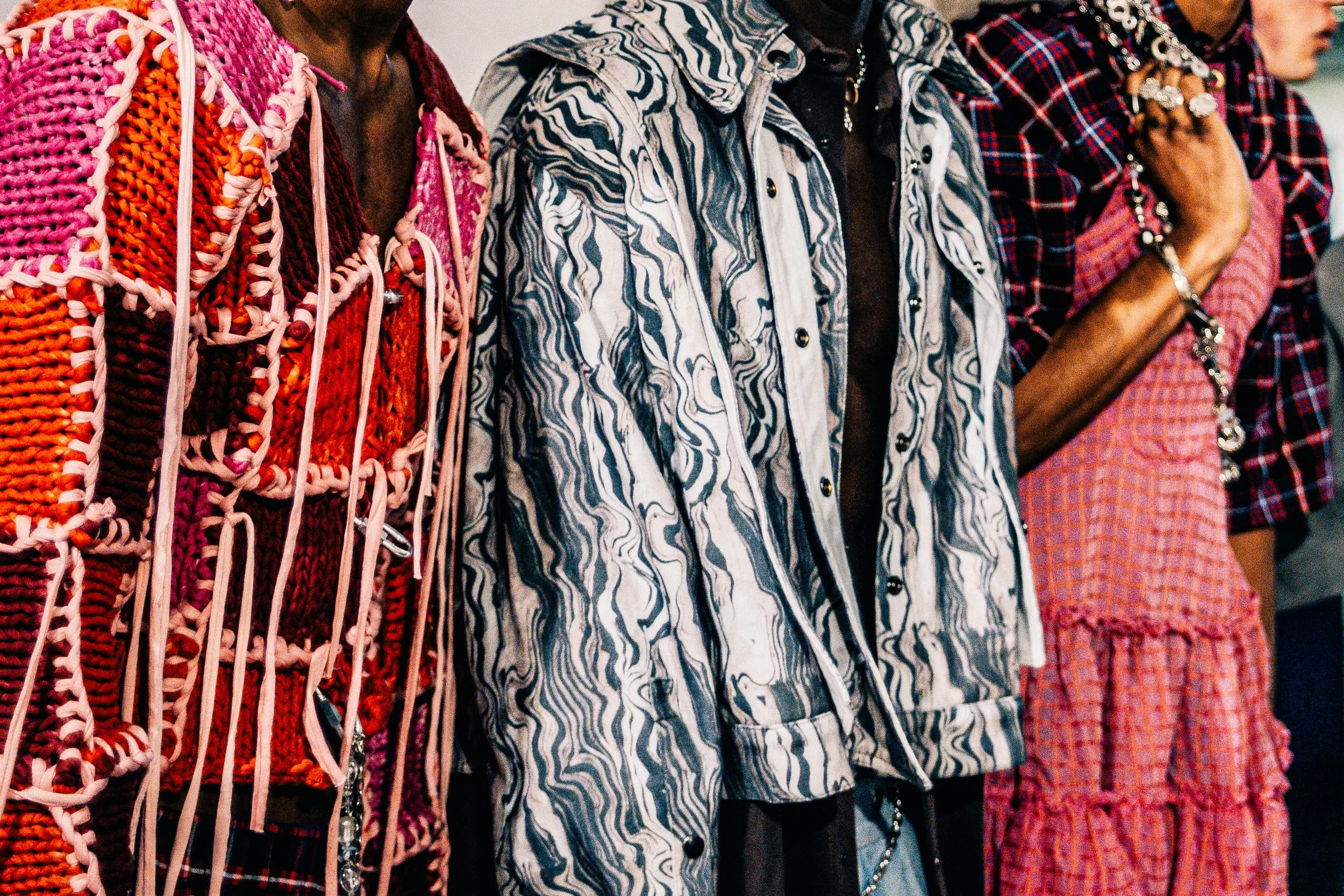 Várias pessoas usando roupas de estampas e cores diferentes, vestindo blusas de diferentes estilos, incluindo uma blusa de padrão listrado preto e branco, uma blusa de xadrez vermelha e preta, e uma blusa de tricô em tons de rosa e laranja, em uma fi
