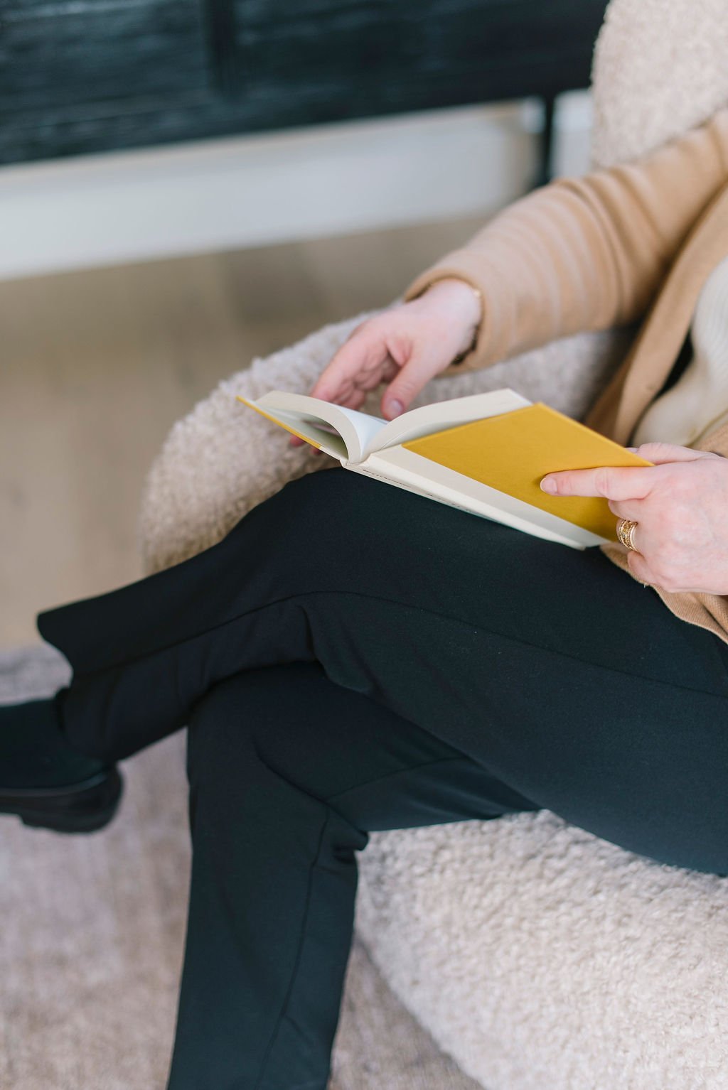 Person sitting on a beige textured couch, holding an open book with a yellow cover. The person is wearing black pants and a beige and white layered top.