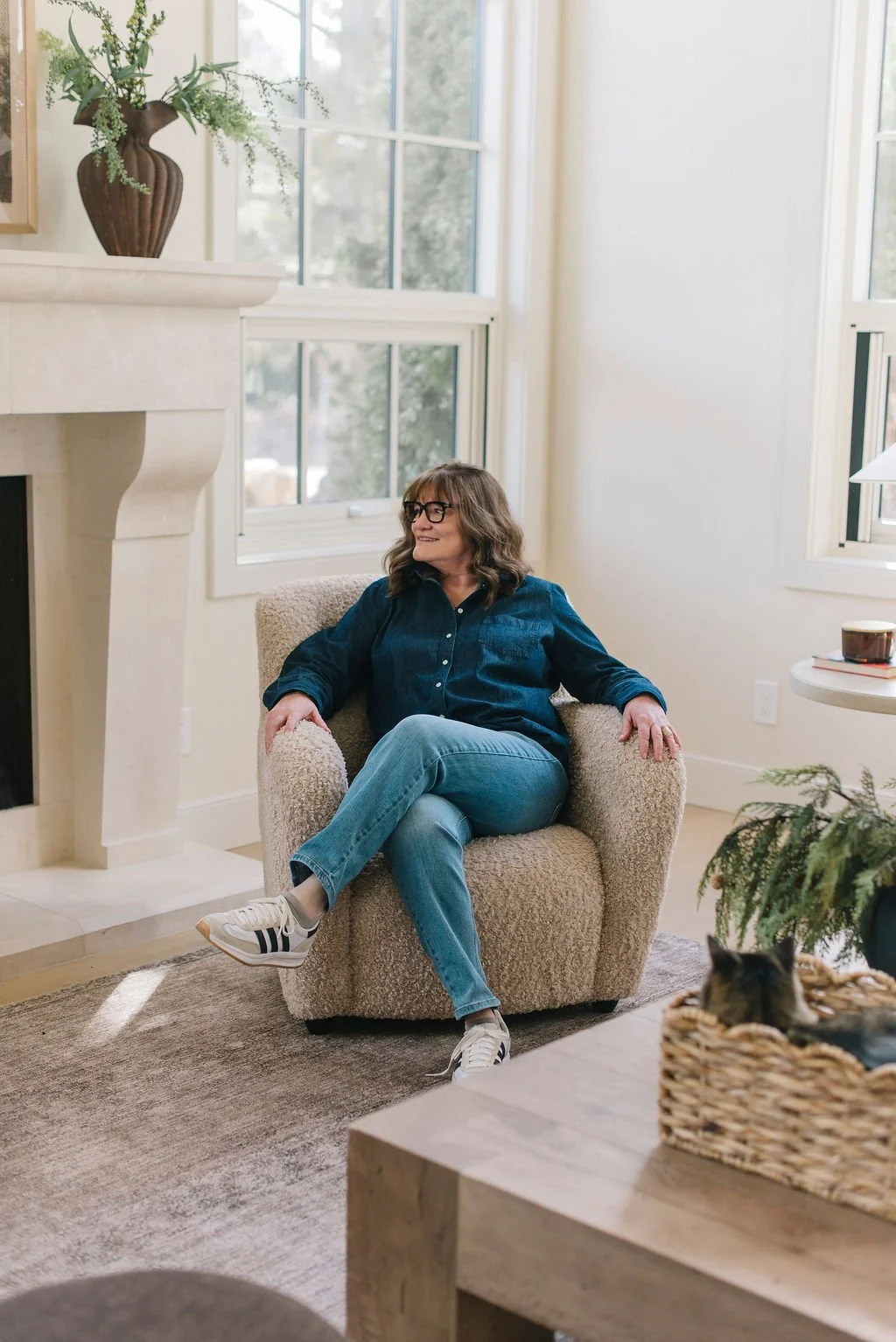 A woman with shoulder-length brown hair, glasses, wearing a denim shirt and jeans, sitting comfortably in a beige armchair in a brightly lit living room with large windows, a fireplace, and a decorative vase with flowers on a mantel. There is a cat in a woven basket on a table nearby.