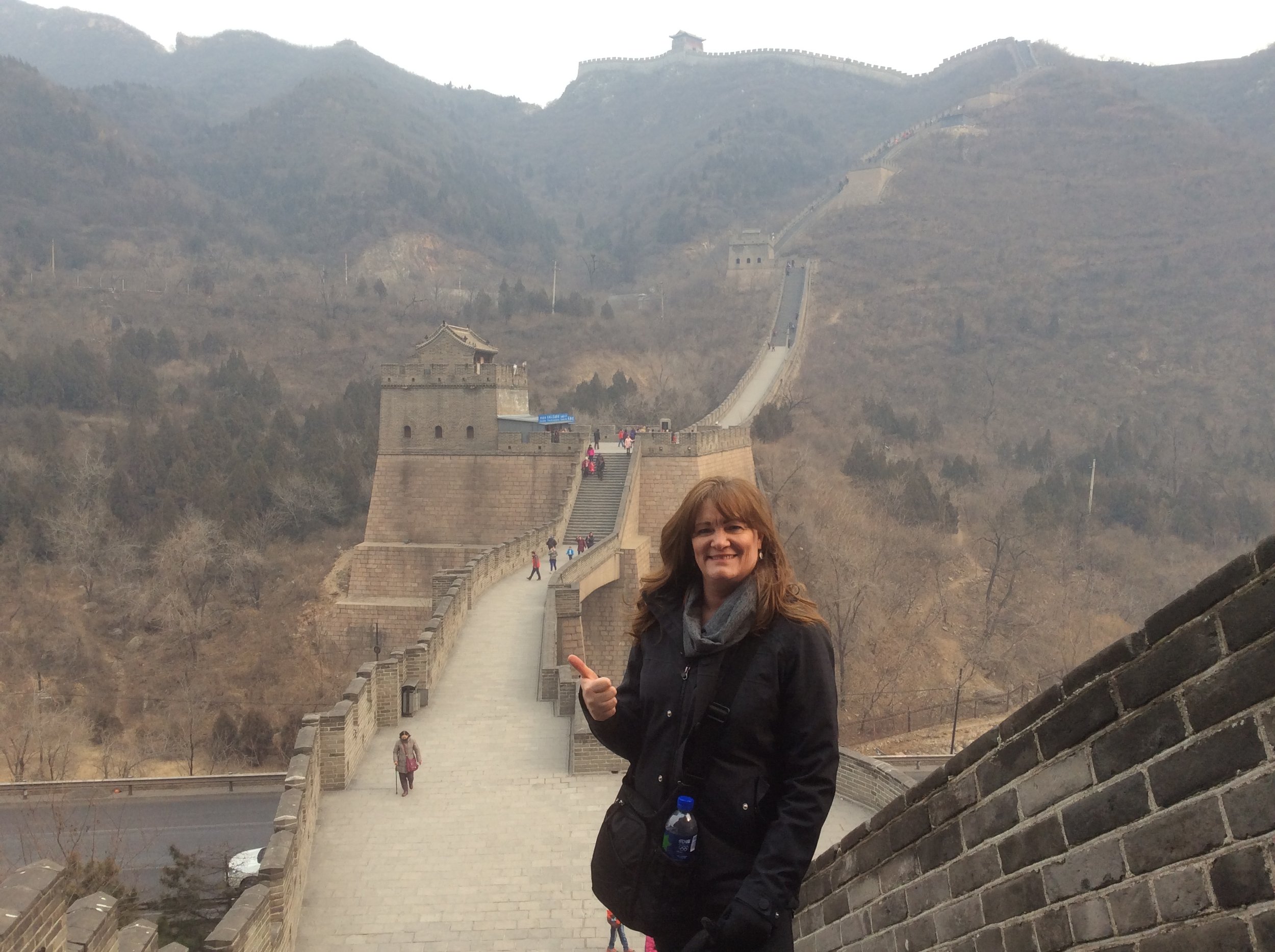 A woman with brown hair, wearing a black jacket and gray scarf, smiling and giving a thumbs-up, standing on the Great Wall of China with mountains and watchtowers in the background.