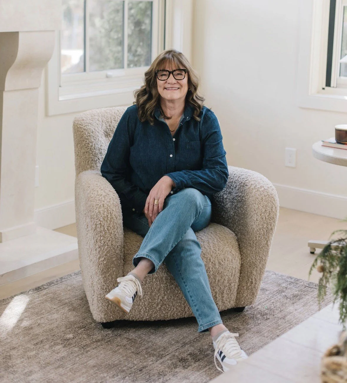 A woman with glasses and shoulder-length hair sitting on a plush beige armchair in a well-lit room with large windows, wearing a denim shirt, jeans, and white sneakers, smiling at the camera.