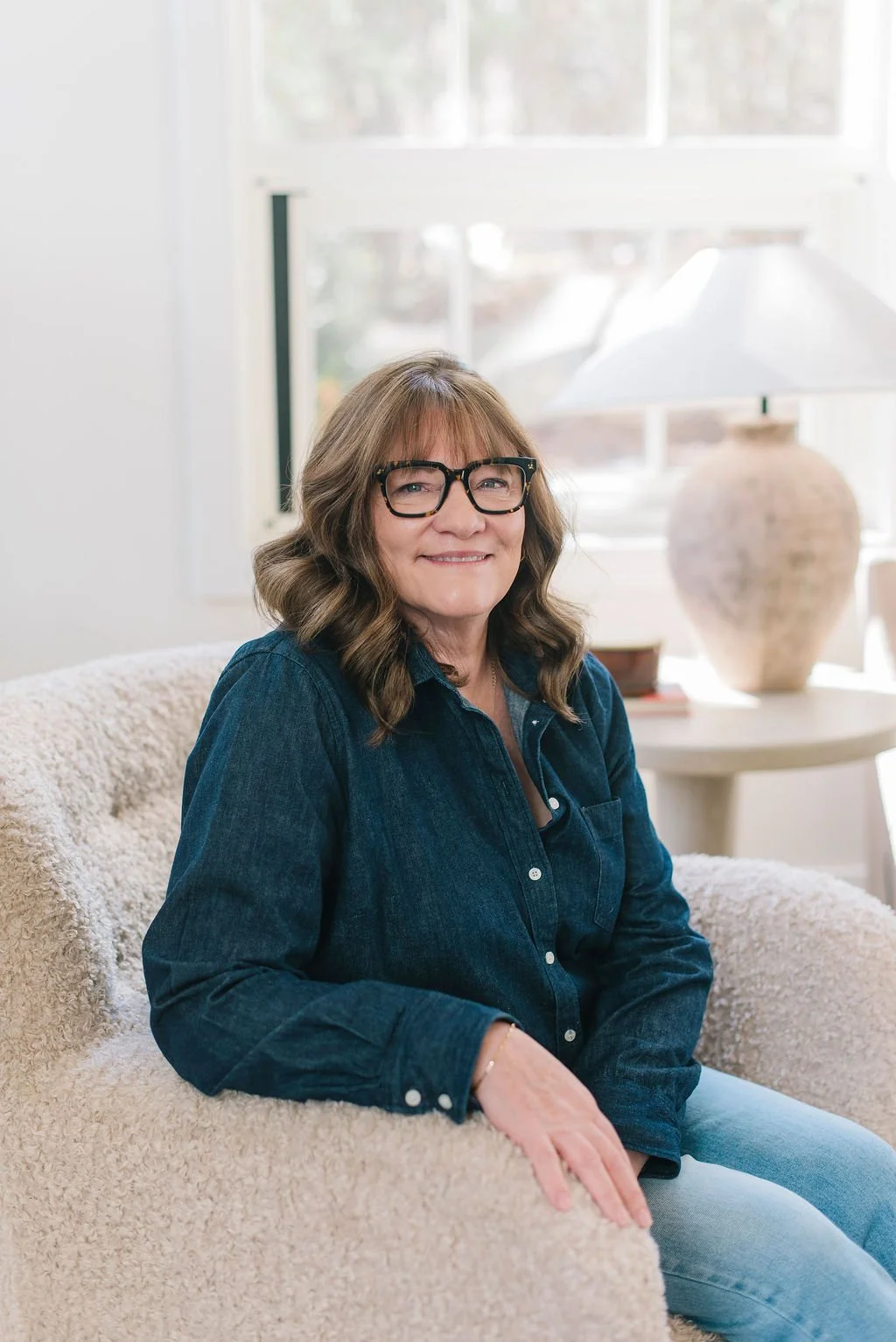 A woman with glasses and wavy brown hair smiling, sitting on a beige couch in a bright room with a large window and a lamp on a table behind her.