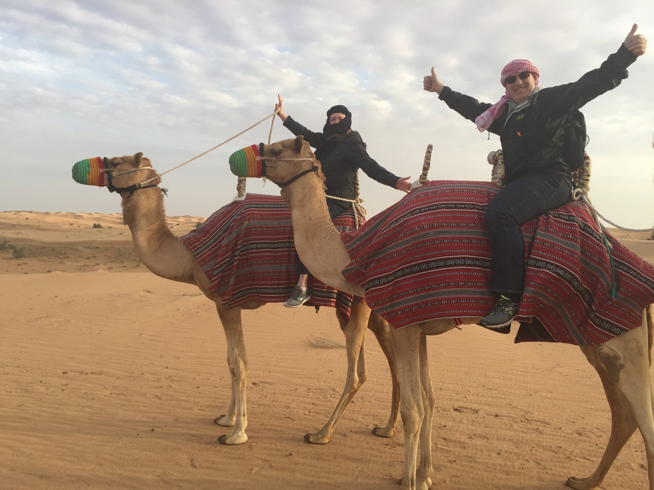 Two women riding camels in a desert landscape, with one woman giving a thumbs up and both enjoying the ride.