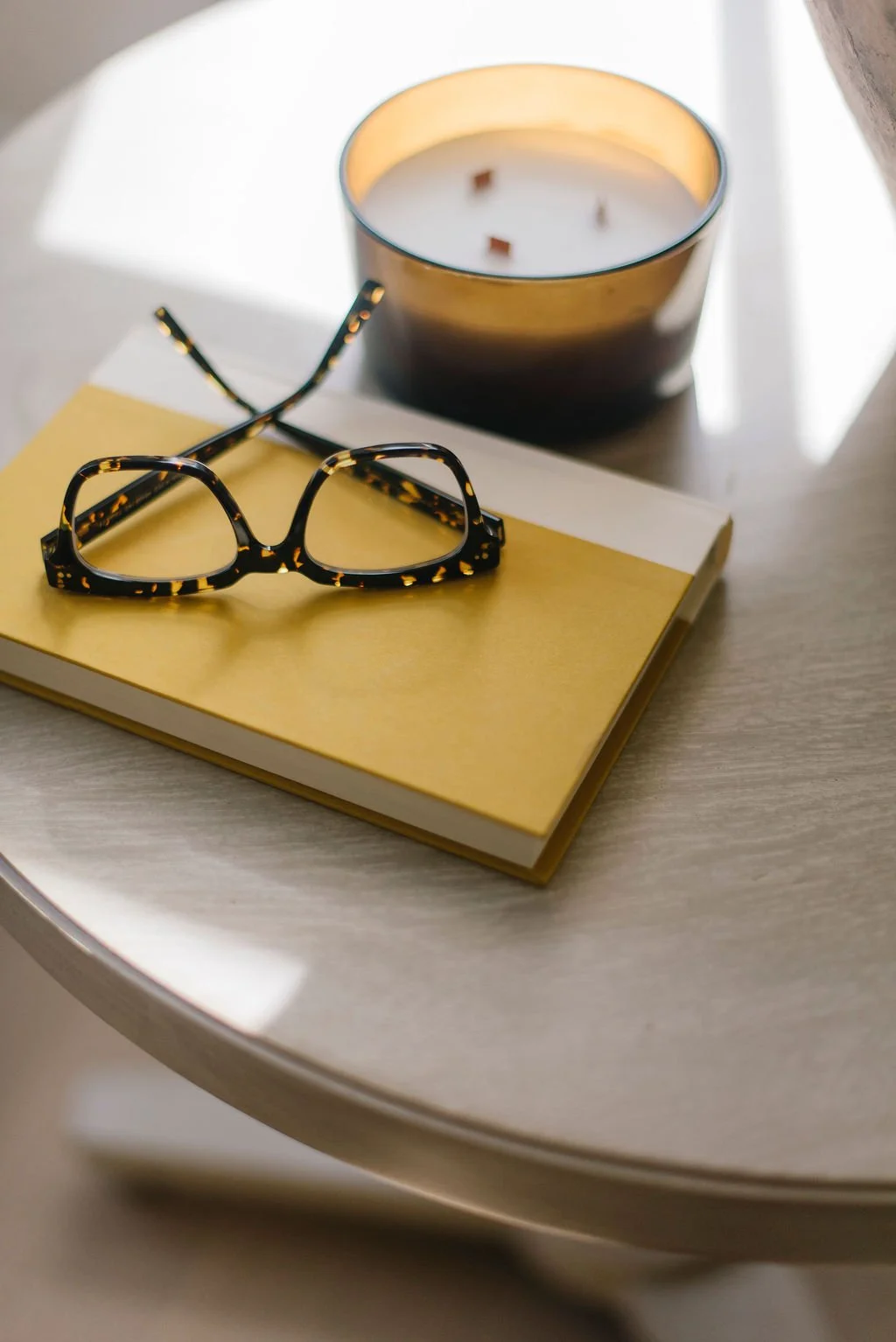 A pair of tortoise-shell glasses resting on a yellow book, with a lit candle in a dark glass holder behind and to the right, on a round white table.