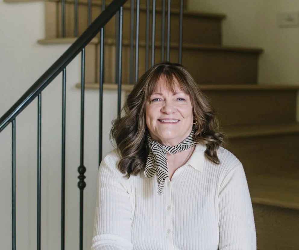 A smiling woman with brown hair and bangs, wearing a white blouse and a black and white striped scarf, sitting on stairs with a black railing in a home interior.
