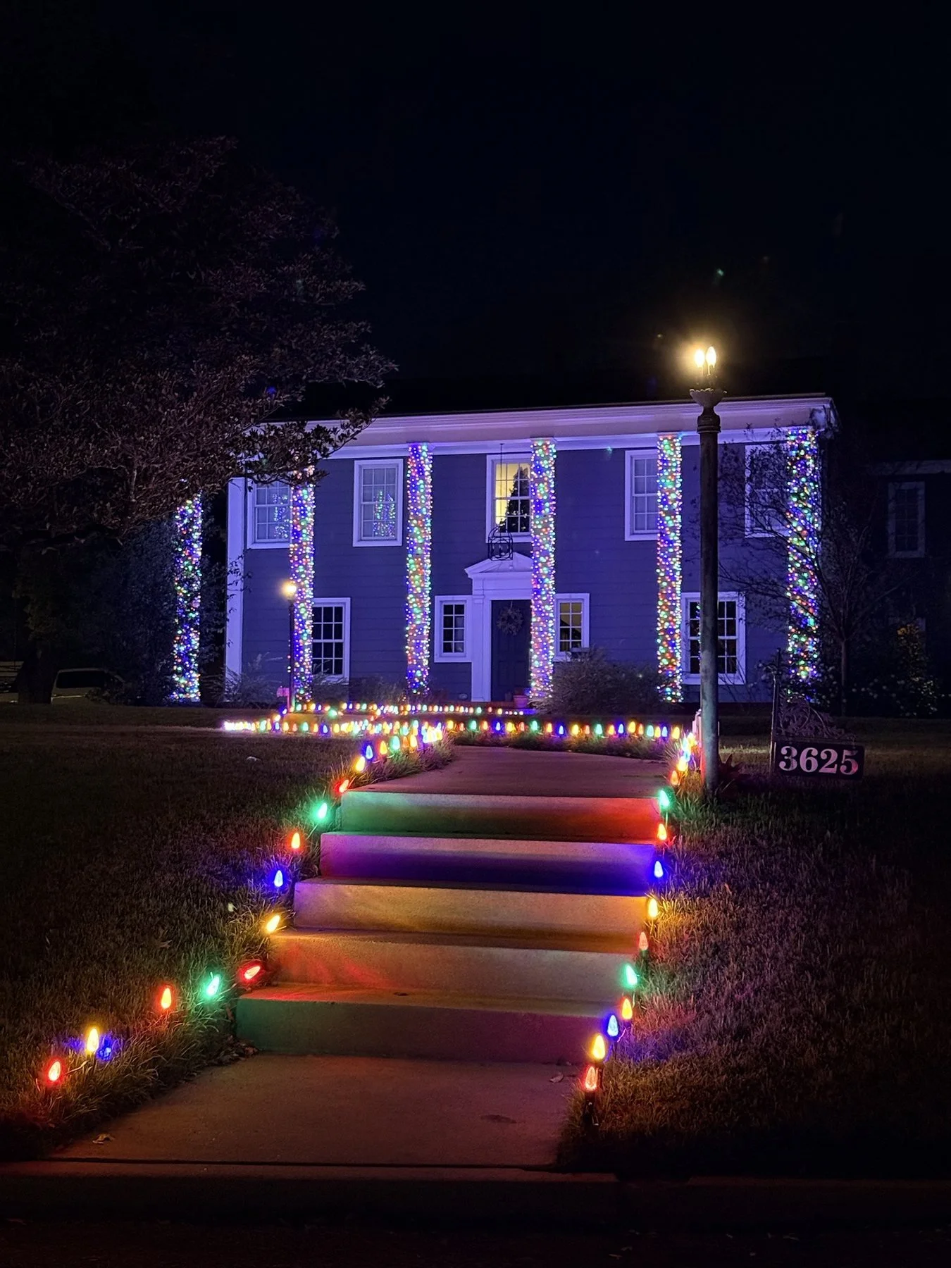 Blue two-story home with multicolor lights wrapped around the columns and a colorful lit walkway