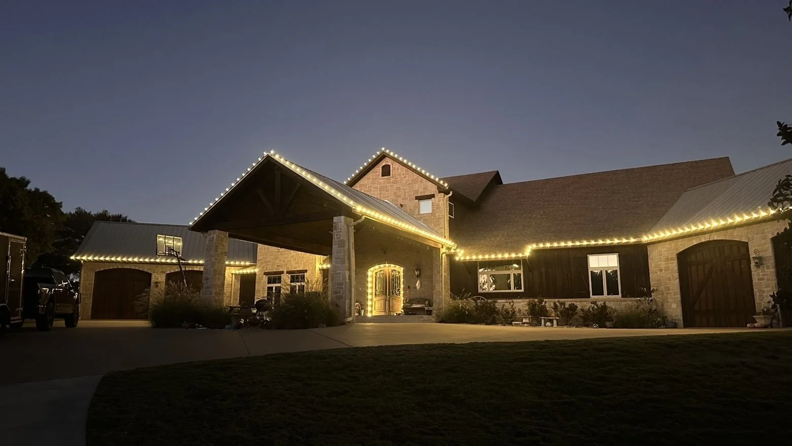 Stone and wood home with warm white lights along the roofline and garage