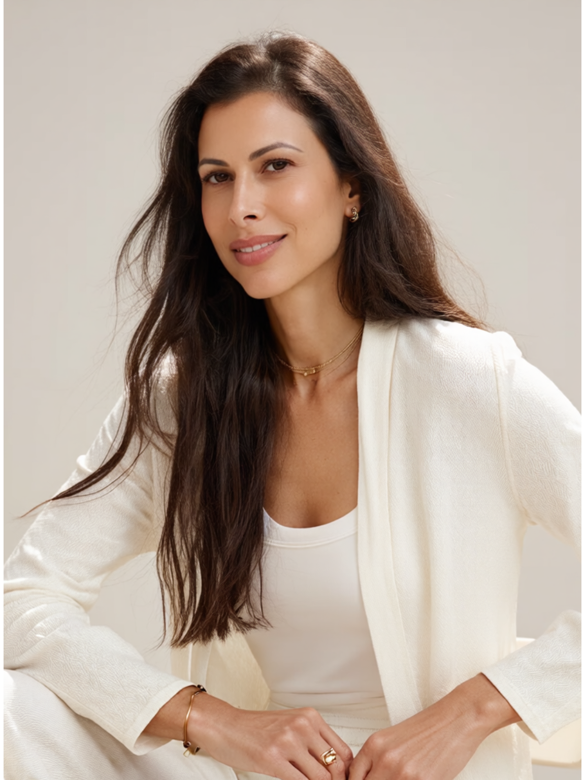 A woman with long dark brown hair and light skin, smiling, wearing a white blazer, white top, and gold jewelry, sitting against a plain light background.