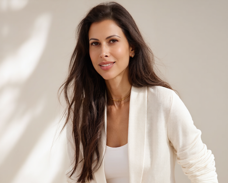 A woman with long brown hair, smiling, wearing a white blazer and a gold necklace against a light background.