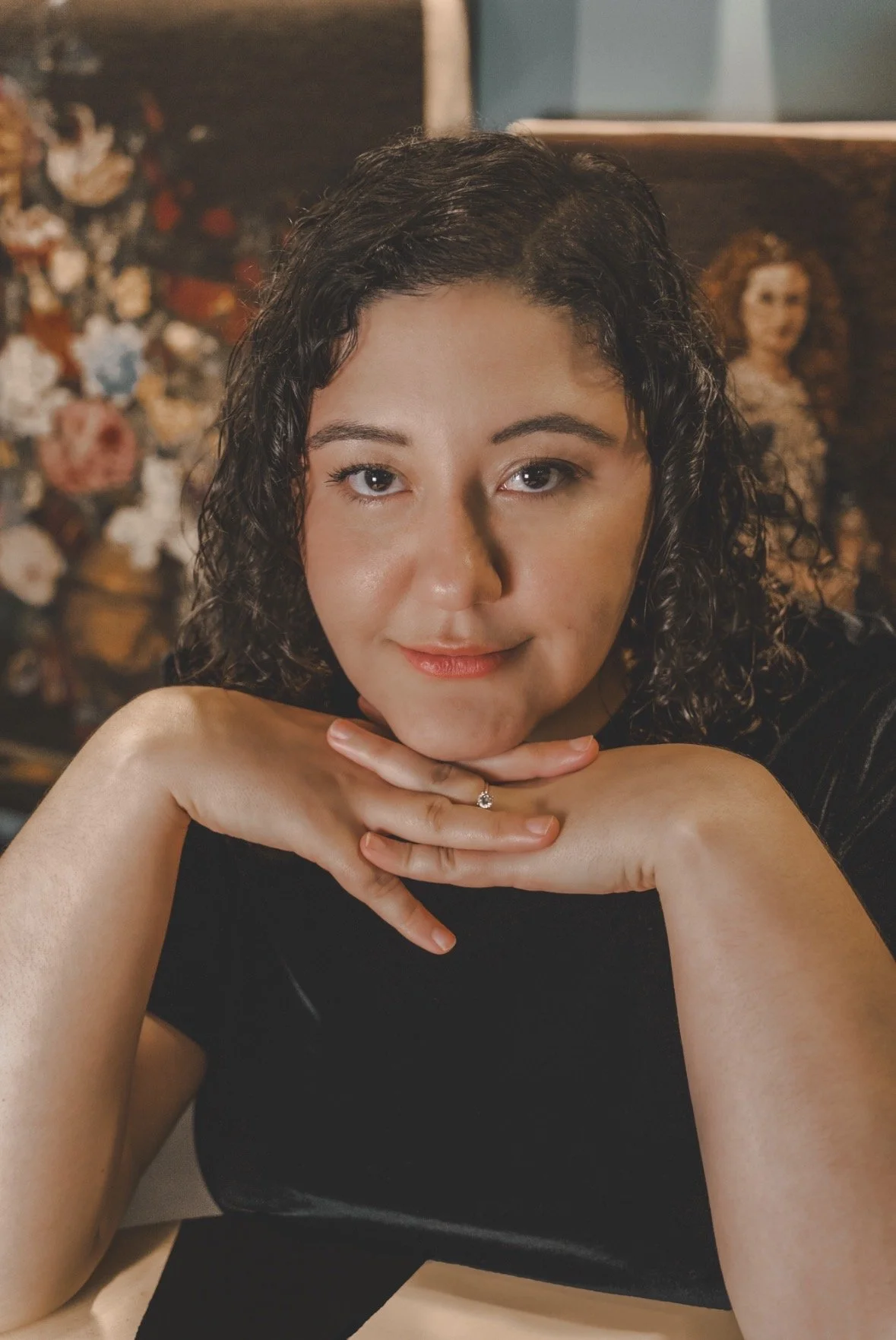 Portrait of Arianna Crawford: dark curly hair, light smile, direct eye contact, head resting on her interlocked hands. She is in front of floral embroidered artwork.