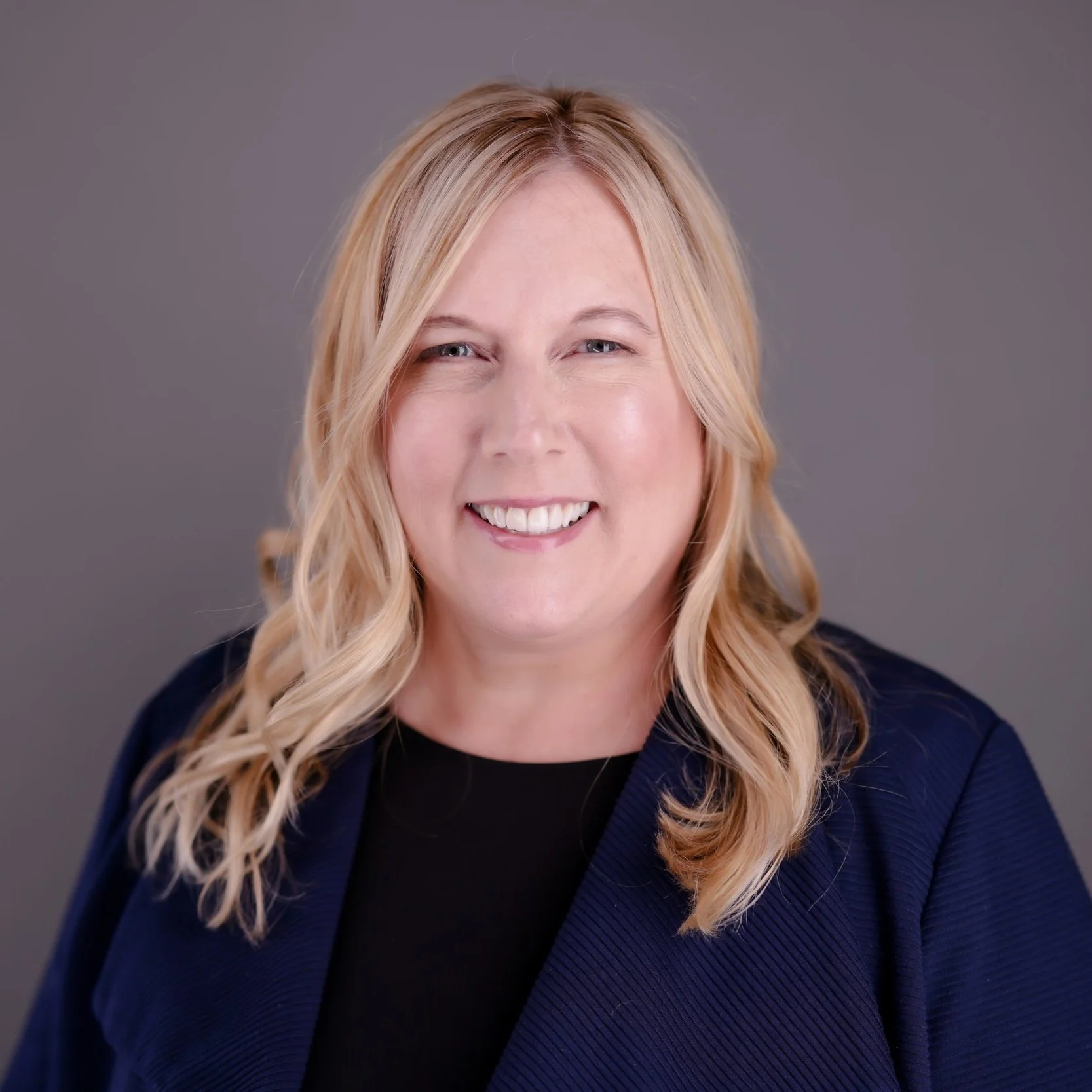A woman with blonde wavy hair smiling at the camera, wearing a black top and a navy blue blazer against a gray background.