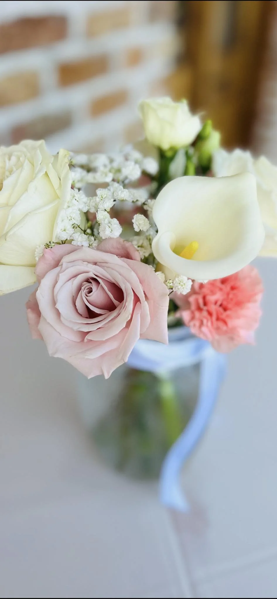 A bouquet of light pink roses, white calla lilies, and small white filler flowers in a glass vase with a light blue ribbon.