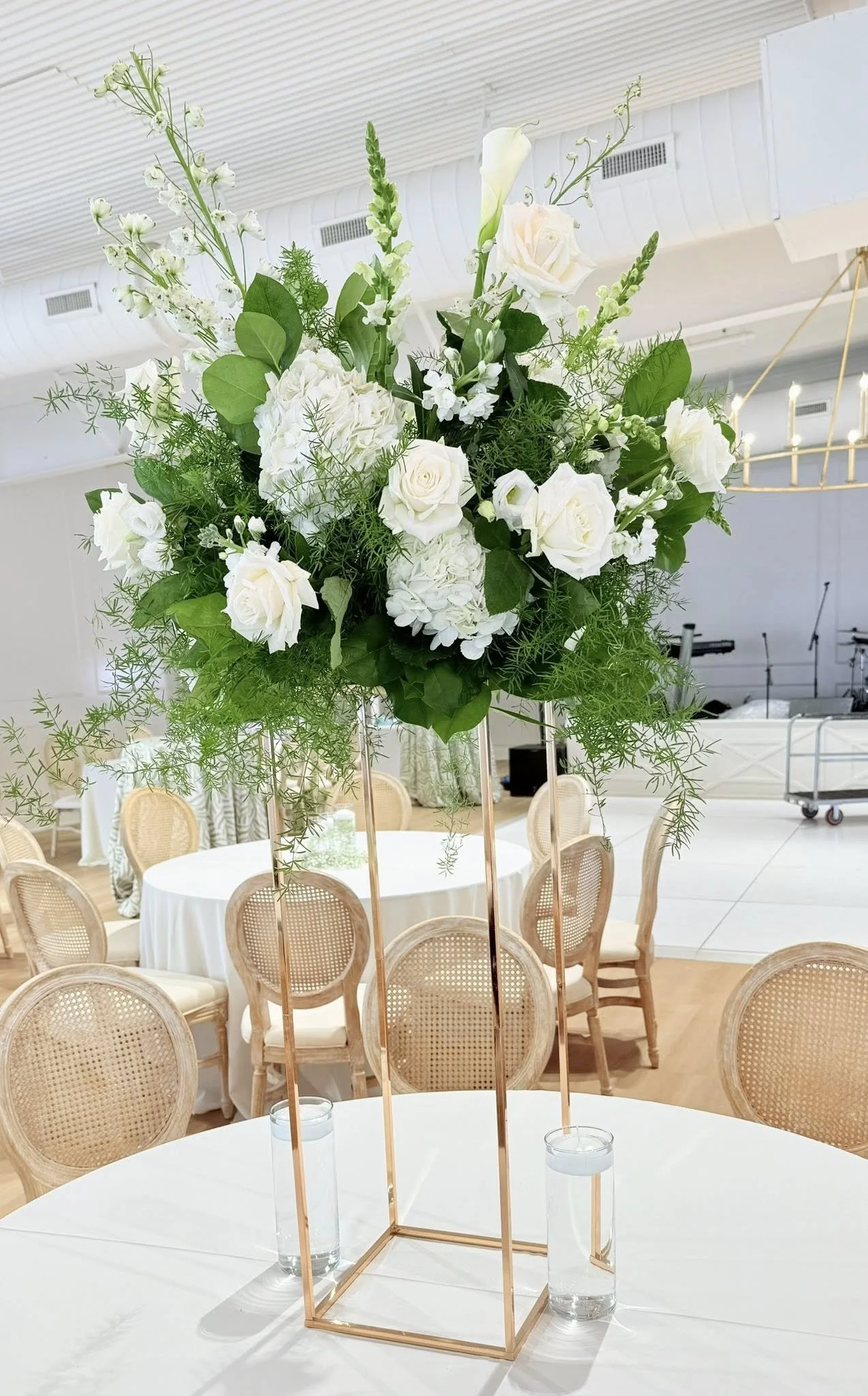 A tall floral arrangement with white roses, hydrangeas, and various greenery in a gold geometric stand, set on a white table at an event venue.