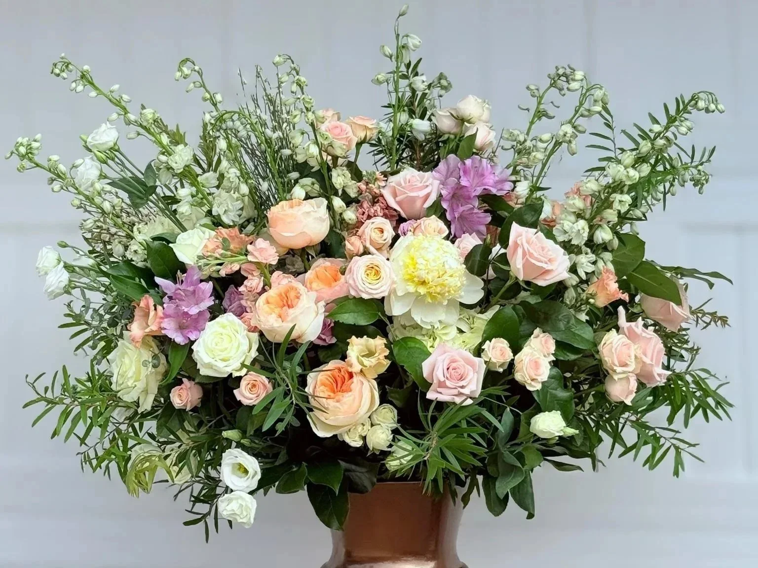 A large flower arrangement in a brown vase featuring pink, white, peach roses, purple lilacs, and greenery.