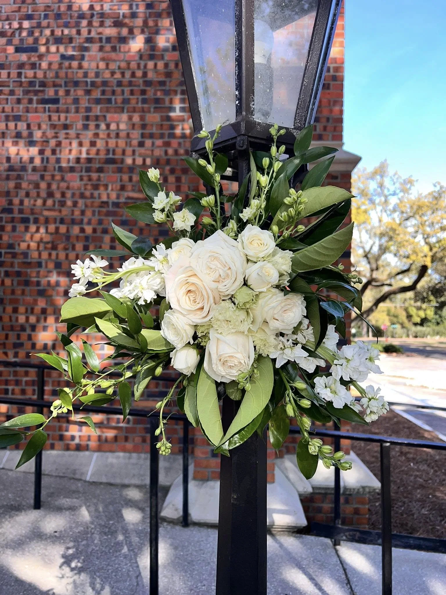 A decorative floral arrangement with white roses, white lisianthus, and greenery attached to a black metal outdoor lamp post near a brick building and sidewalk.