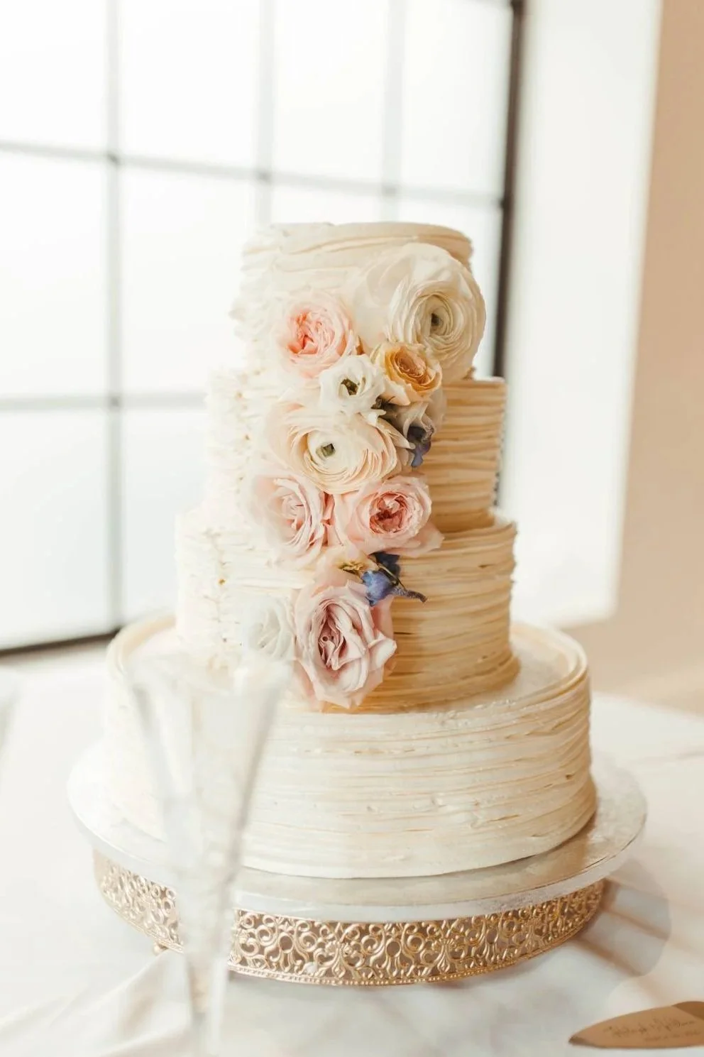 Elegant wedding cake with two tiers, decorated with pastel-colored roses and ruffled frosting, placed on a gold ornate stand near a window.