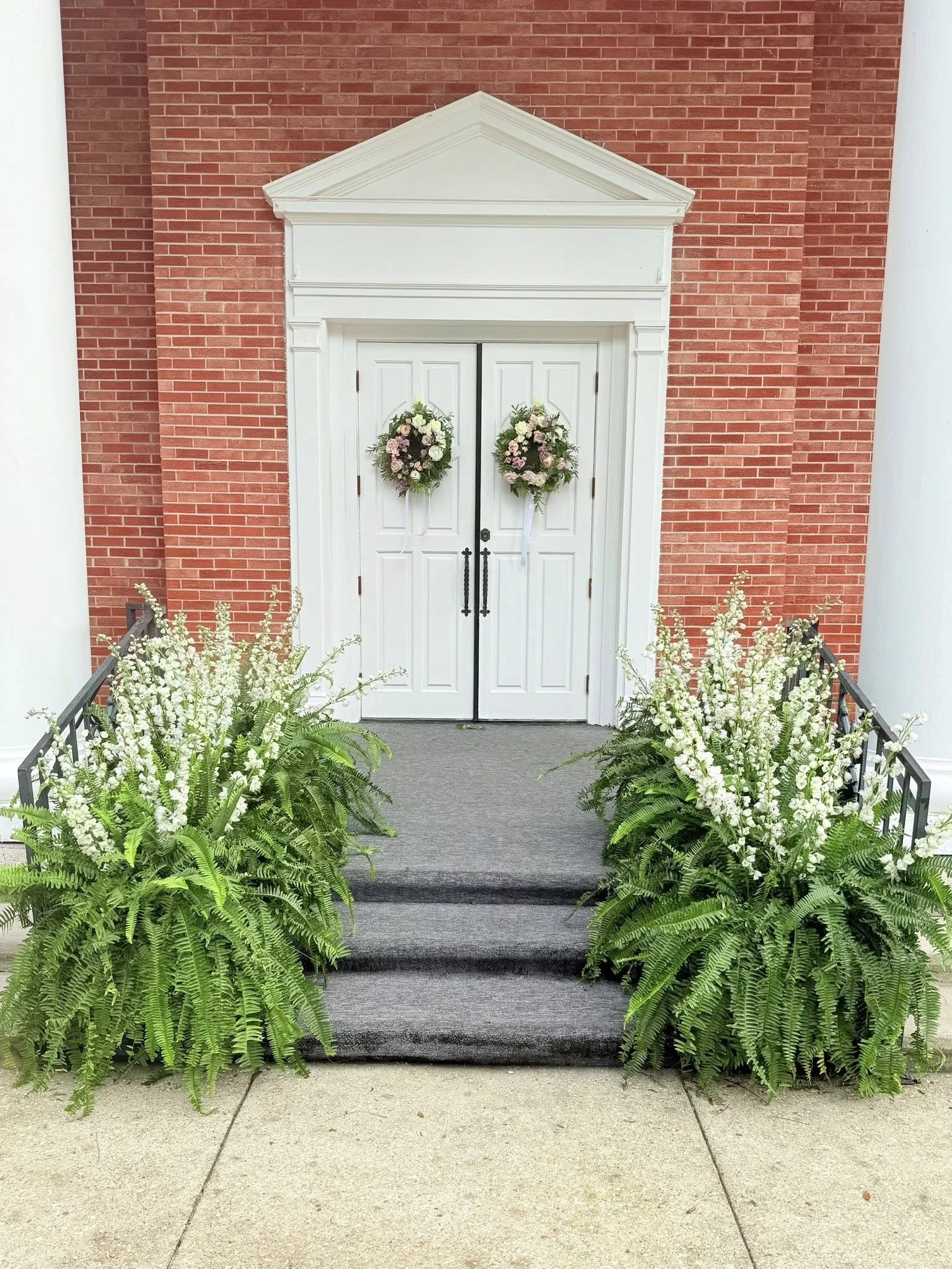 White double doors with floral wreaths, floral arrangements and ferns on a gray carpeted ramp leading to entrance of brick building.
