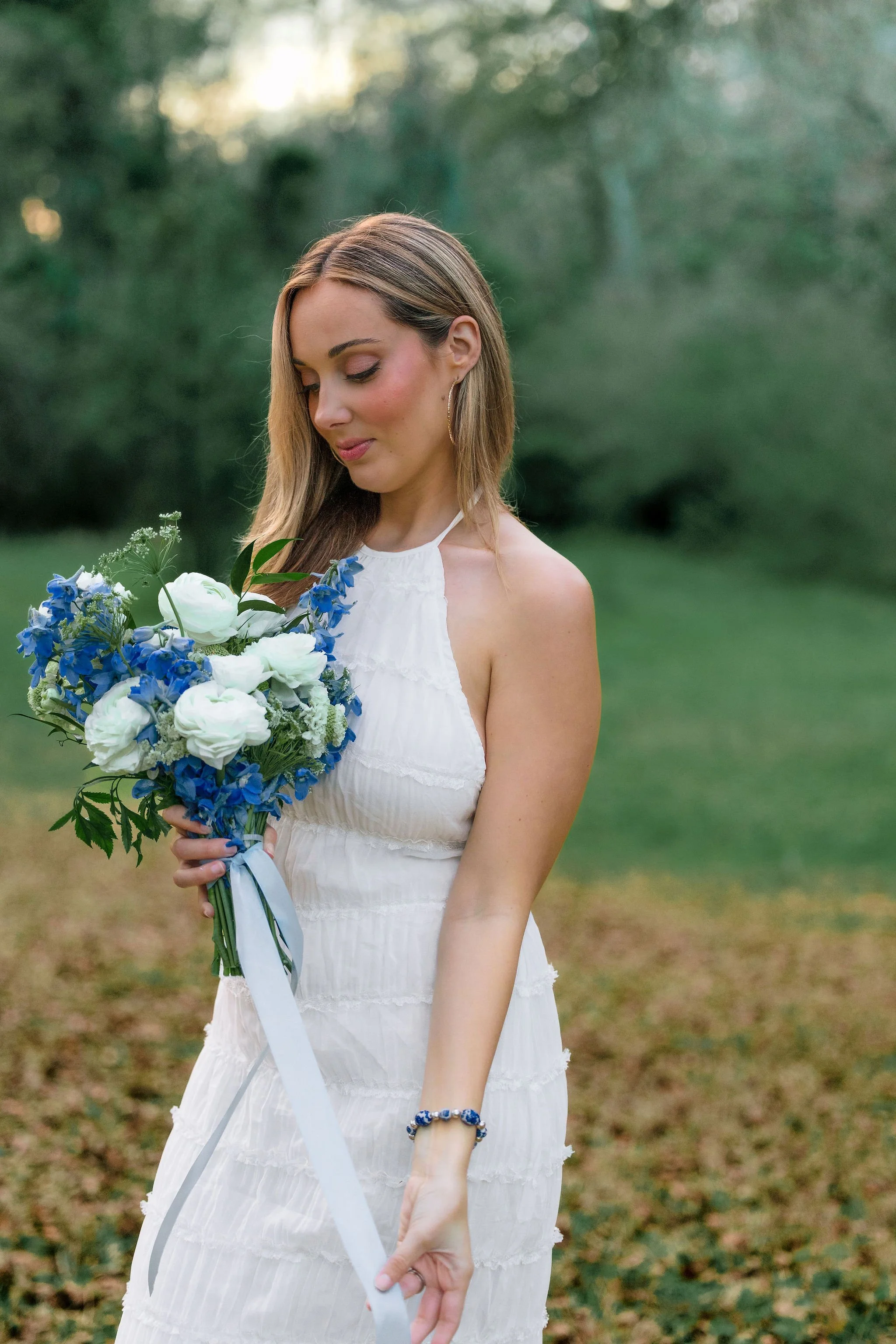 A woman in a white dress holding a bouquet of white and blue flowers outdoors.