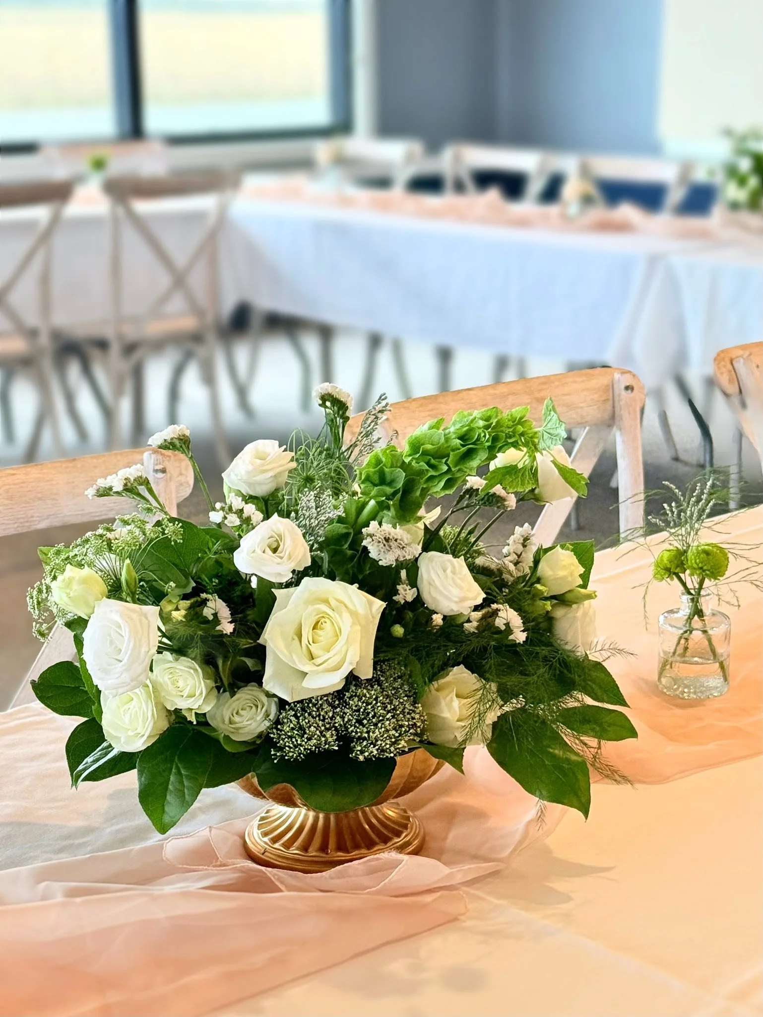 Elegant table setting with a large flower bouquet of white roses and greenery in a gold vase, and small green floral accents in a clear glass vase, on a peach-colored tablecloth.