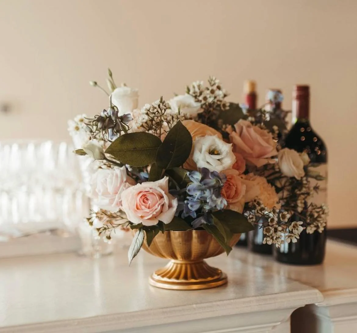 A floral arrangement with pink, white, and purple flowers in a gold vase, placed on a white surface with bottles of wine in the background.