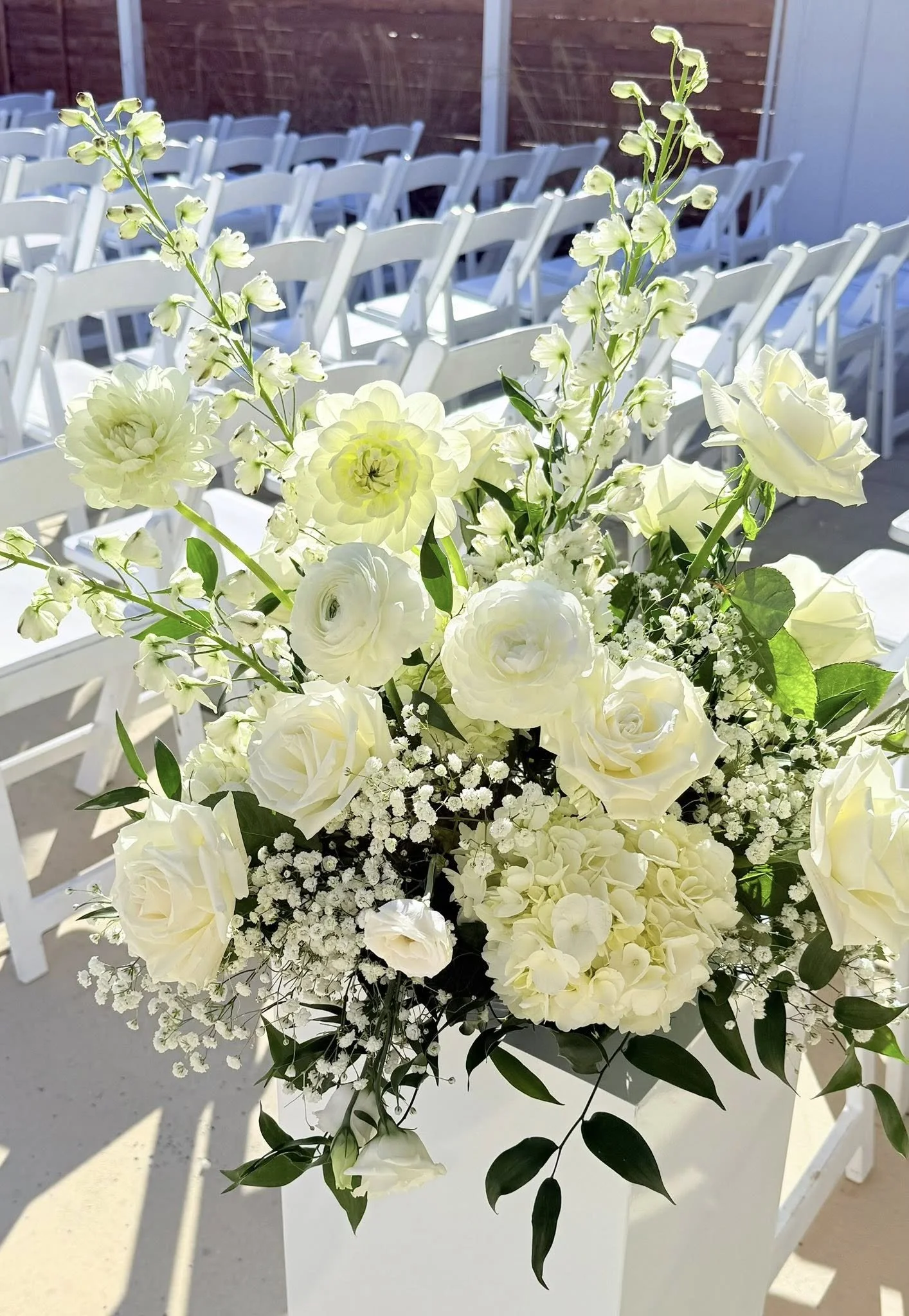 White flower centerpiece with roses, hydrangeas, and baby's breath on a table at an outdoor event, with white chairs in the background.