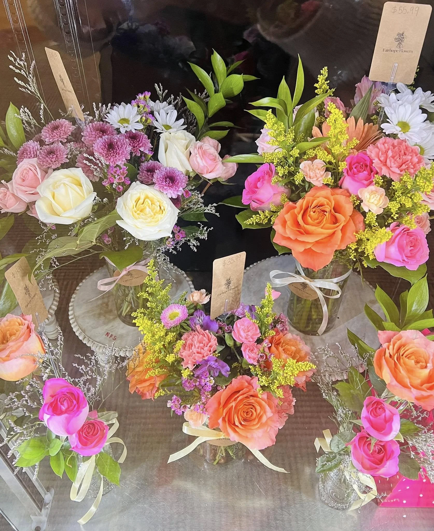 Various colorful flower arrangements including pink, orange, and white roses, daisies, and other flowers displayed in glass vases on a table.