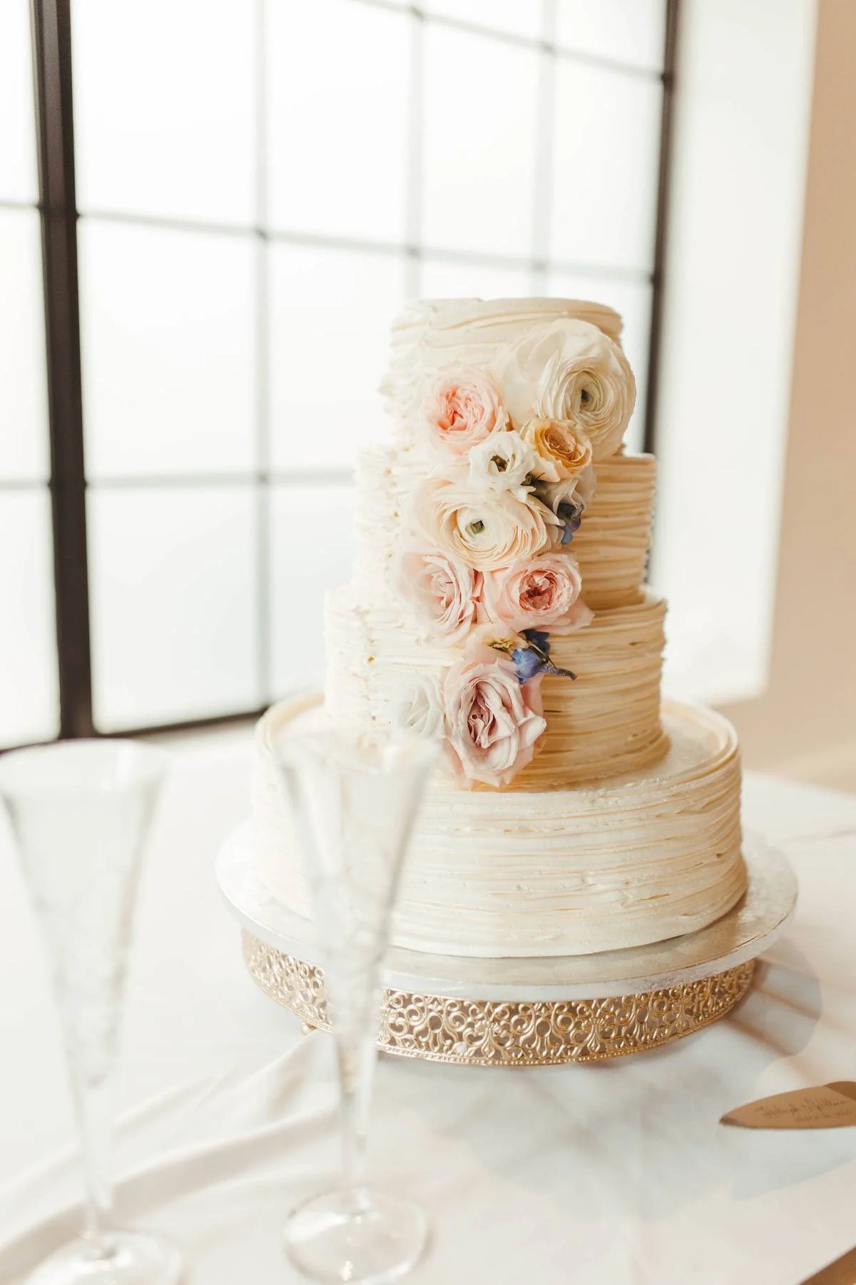 Elegant three-tier wedding cake decorated with pink and white roses and ranunculus, with a textured cream-colored frosting, on a decorative gold cake stand.