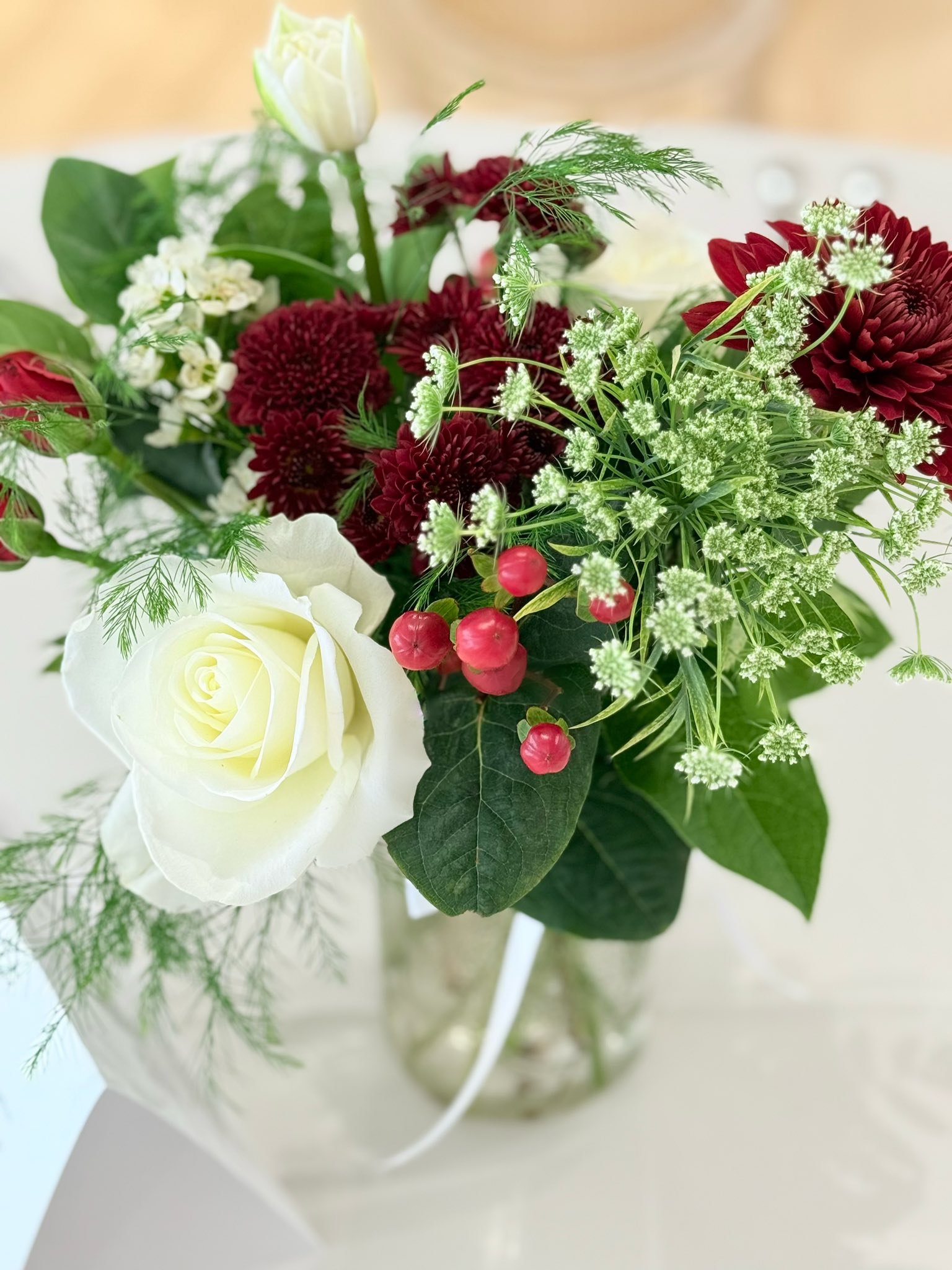 A bouquet of fresh flowers including white roses, red chrysanthemums, small red berries, and white filler flowers, arranged with greenery in a clear glass vase.