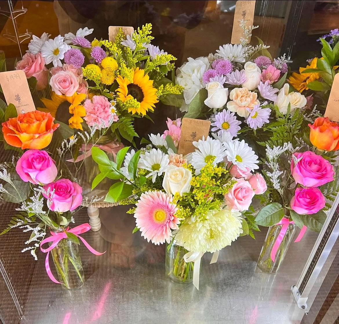 Multiple floral arrangements in glass vases with pink, white, yellow, and purple flowers on a metal surface, each with a small brown price tag attached.