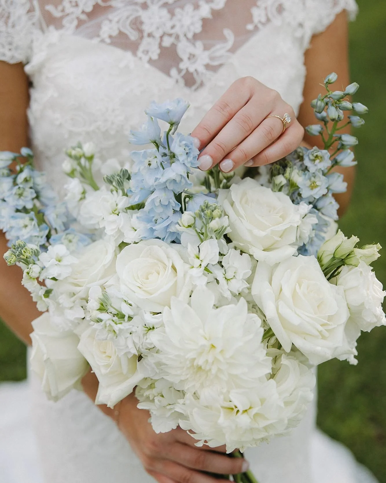 Close-up of a bride holding a bouquet of white and light blue flowers, including roses and delphiniums, with her left hand resting on top, showing a wedding ring.