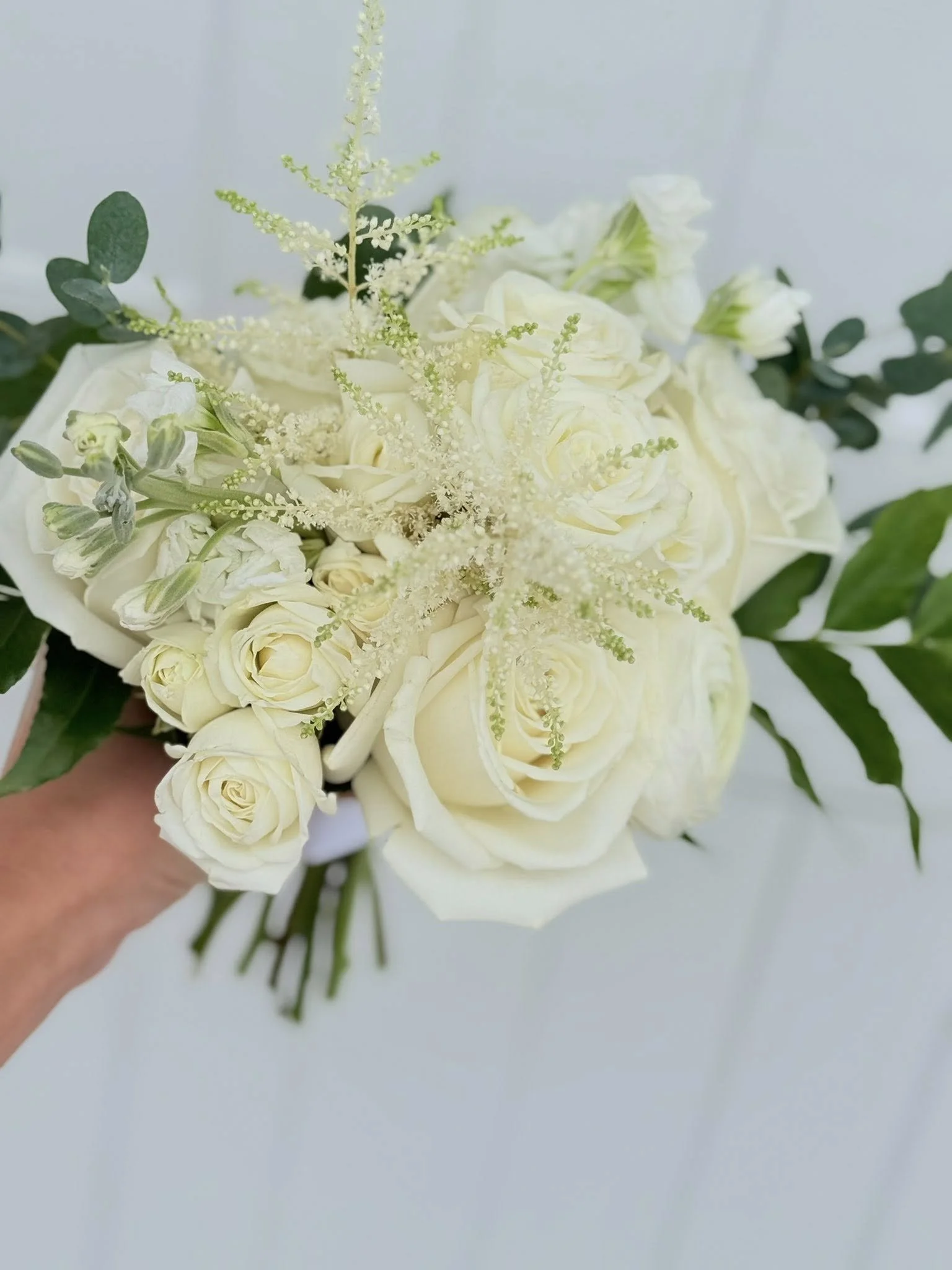 A bouquet of white roses, greenery, and delicate white filler flowers against a blurred light background.