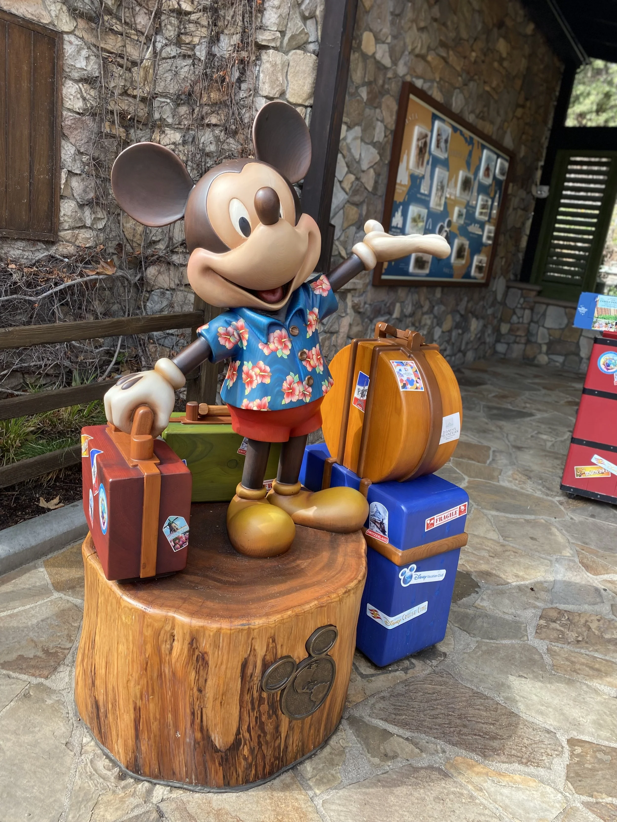 Statue of a cartoon mouse character wearing a floral shirt, standing on a wooden platform with various suitcases. The background is a stone wall with framed pictures.
