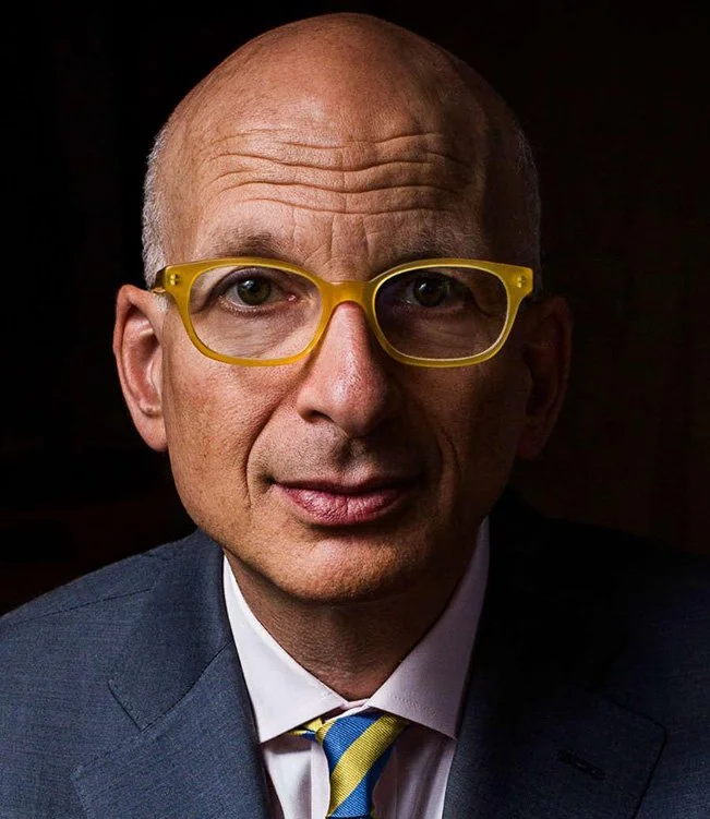 A middle-aged man with a bald head and light skin, wearing yellow glasses, a blue suit, a white shirt, and a blue and yellow striped tie.