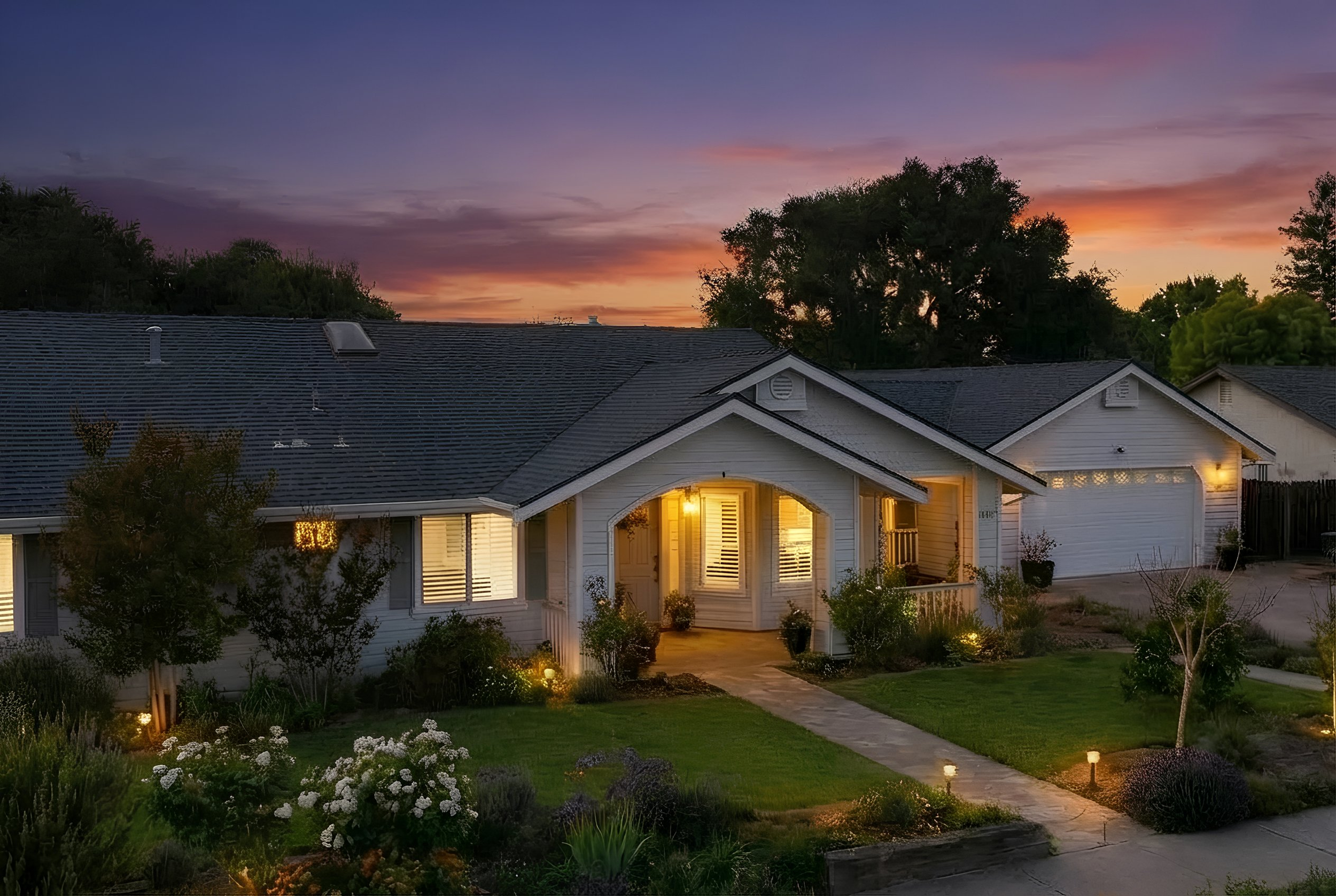 A cozy suburban home at sunset with warm exterior lighting, a well-maintained lawn, and garden, under a colorful sky.