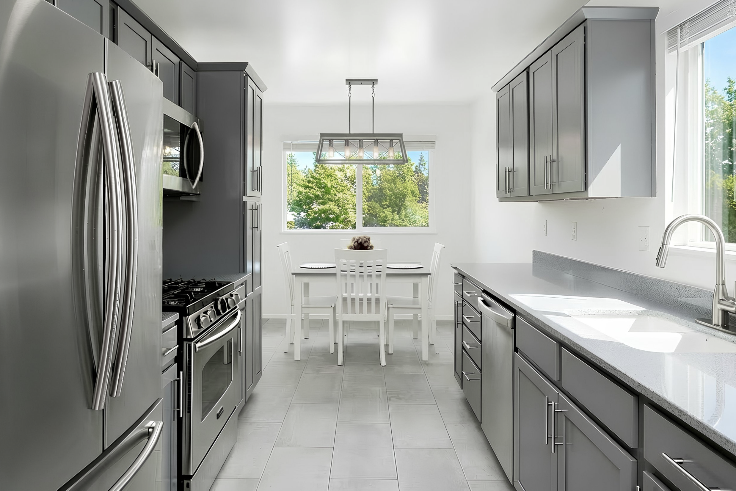 Modern kitchen with gray cabinets, stainless steel appliances, a white dining table with four chairs, and large windows showing green trees outside.