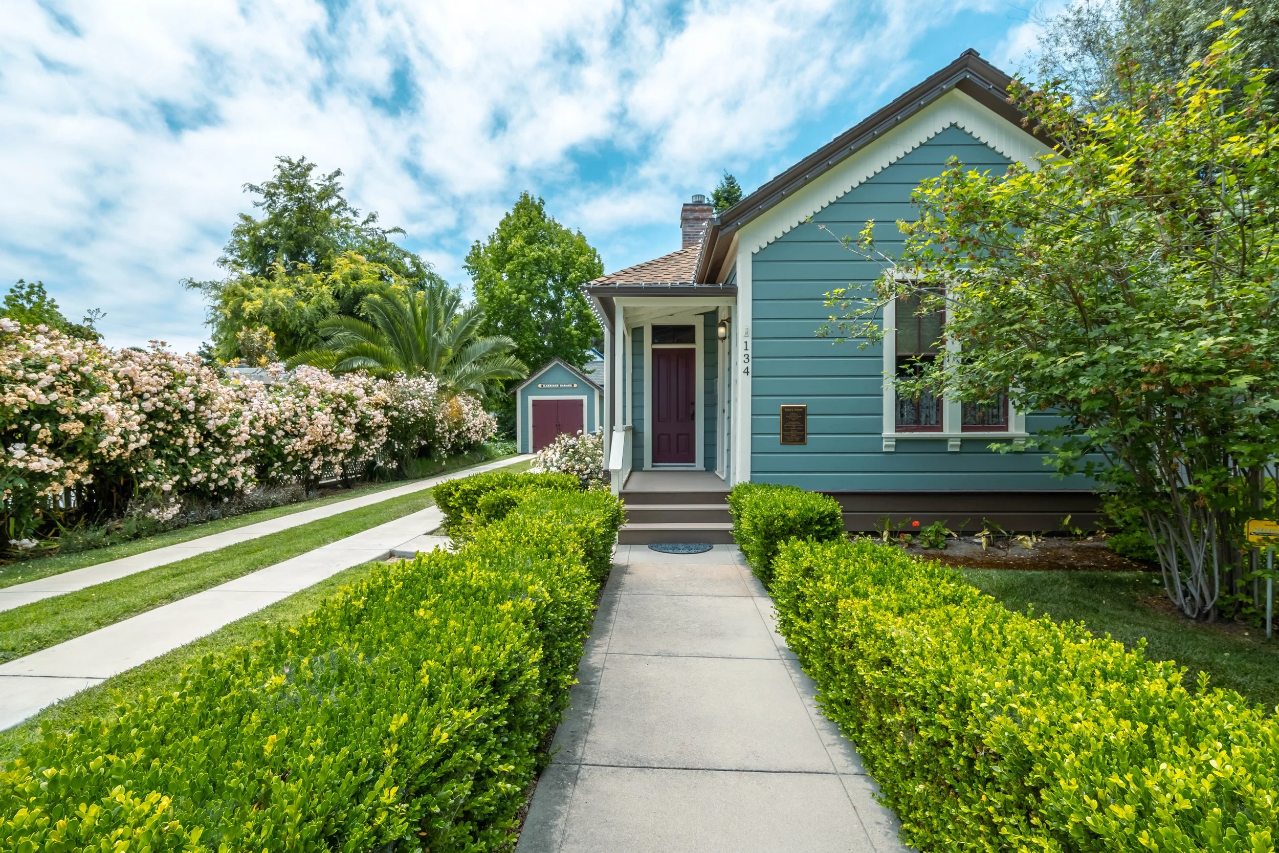 Front view of a blue house with a porch, surrounded by green bushes, trees, and blooming pink flowers under a partly cloudy sky.