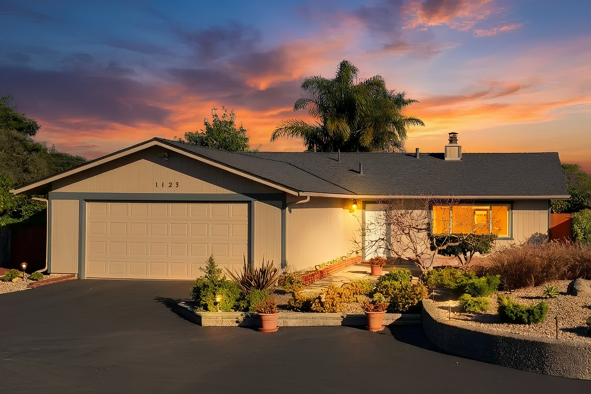 A single-story house with gray siding and a dark roof, illuminated by outdoor lights, at sunset with colorful sky and palm trees in the background.