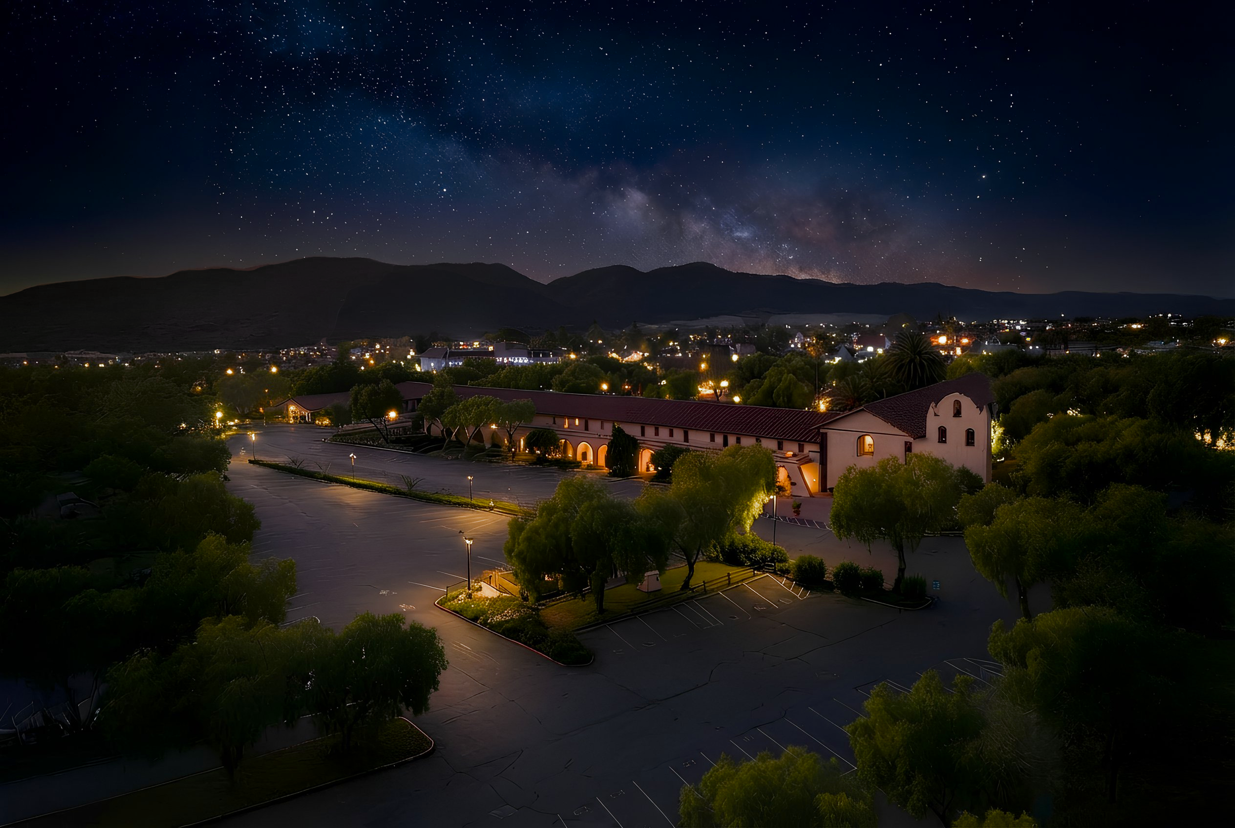 Nighttime view of a parking lot surrounded by trees, with a building that has arched windows and a red-tiled roof. In the background, a cityscape with lights, mountains, and a starry sky with the Milky Way are visible.