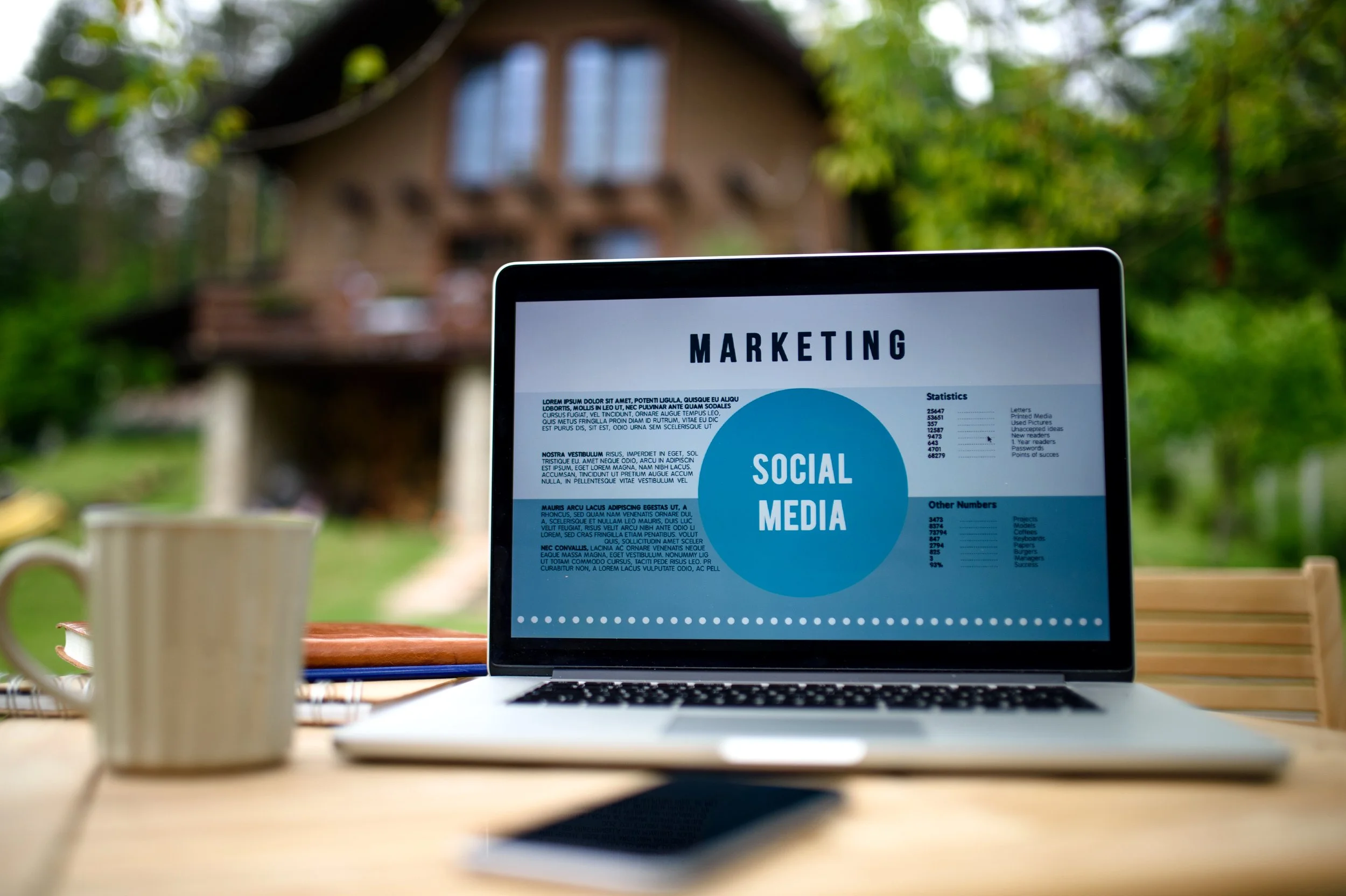 Laptop on a wooden table displaying a marketing presentation about social media, with a mug and notebook nearby, outside in a garden with a house in the background.