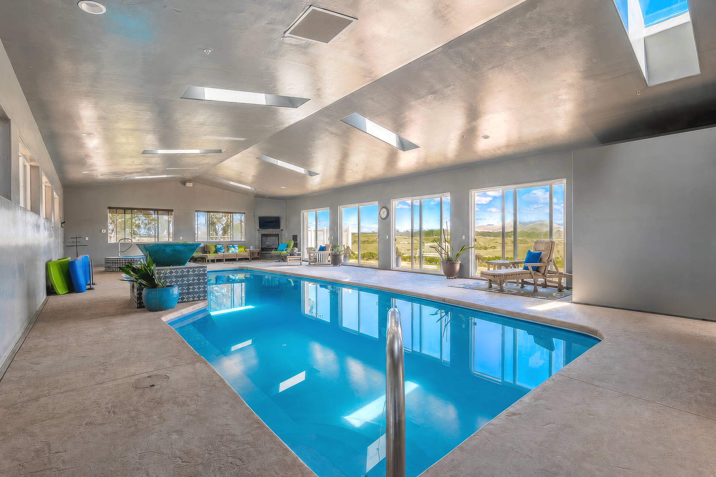 Indoor swimming pool with large windows showing a landscape of hills and blue sky, modern furniture, plants, and a metal pool ladder.