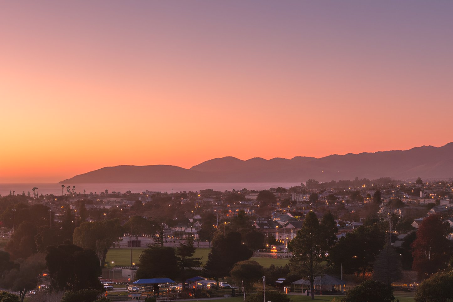 Sunset over a coastal town with mountains in the background, trees in the foreground, and city lights starting to turn on.